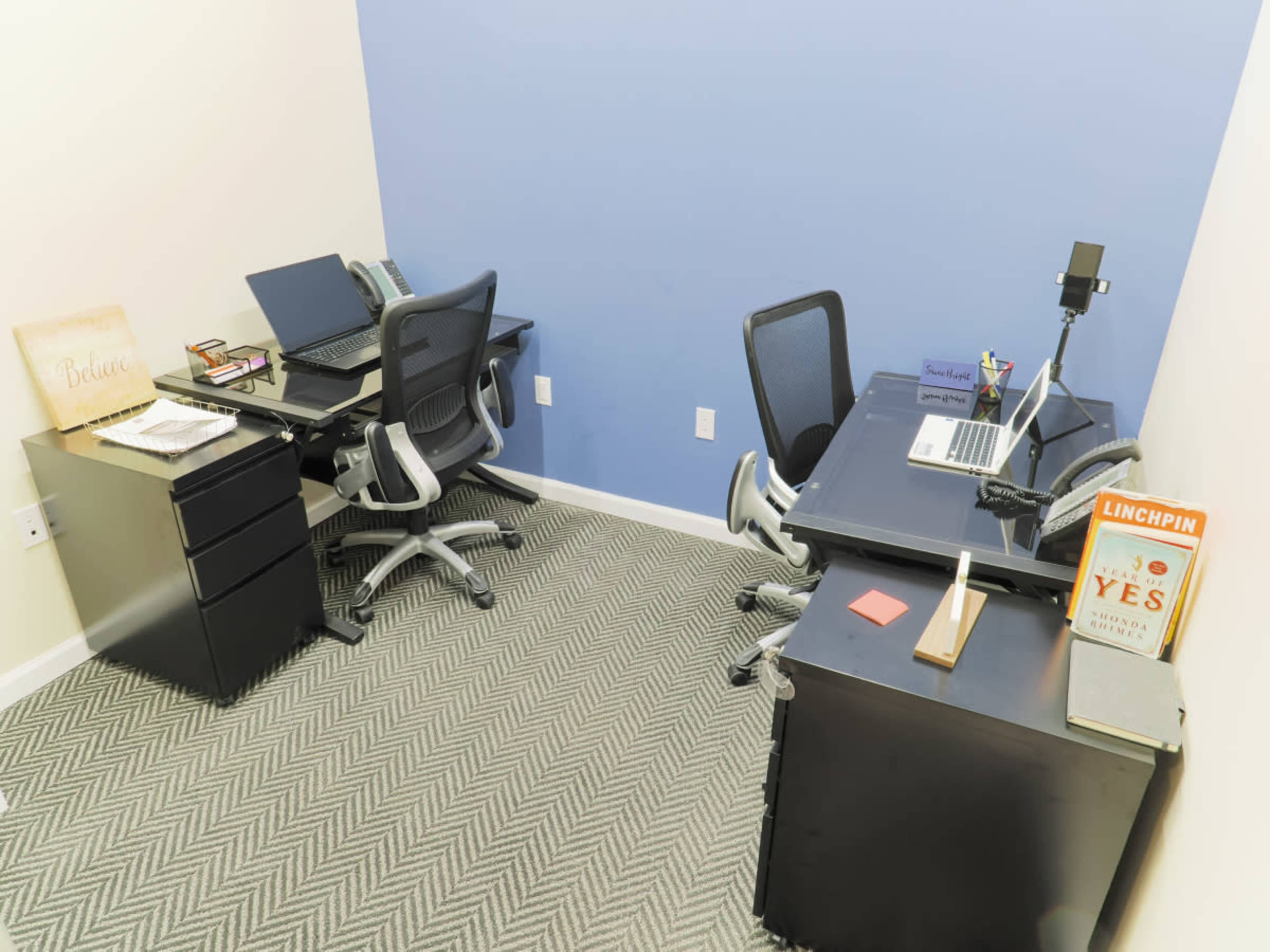 A small office space with two black desks equipped with computers and office supplies against blue and beige walls.