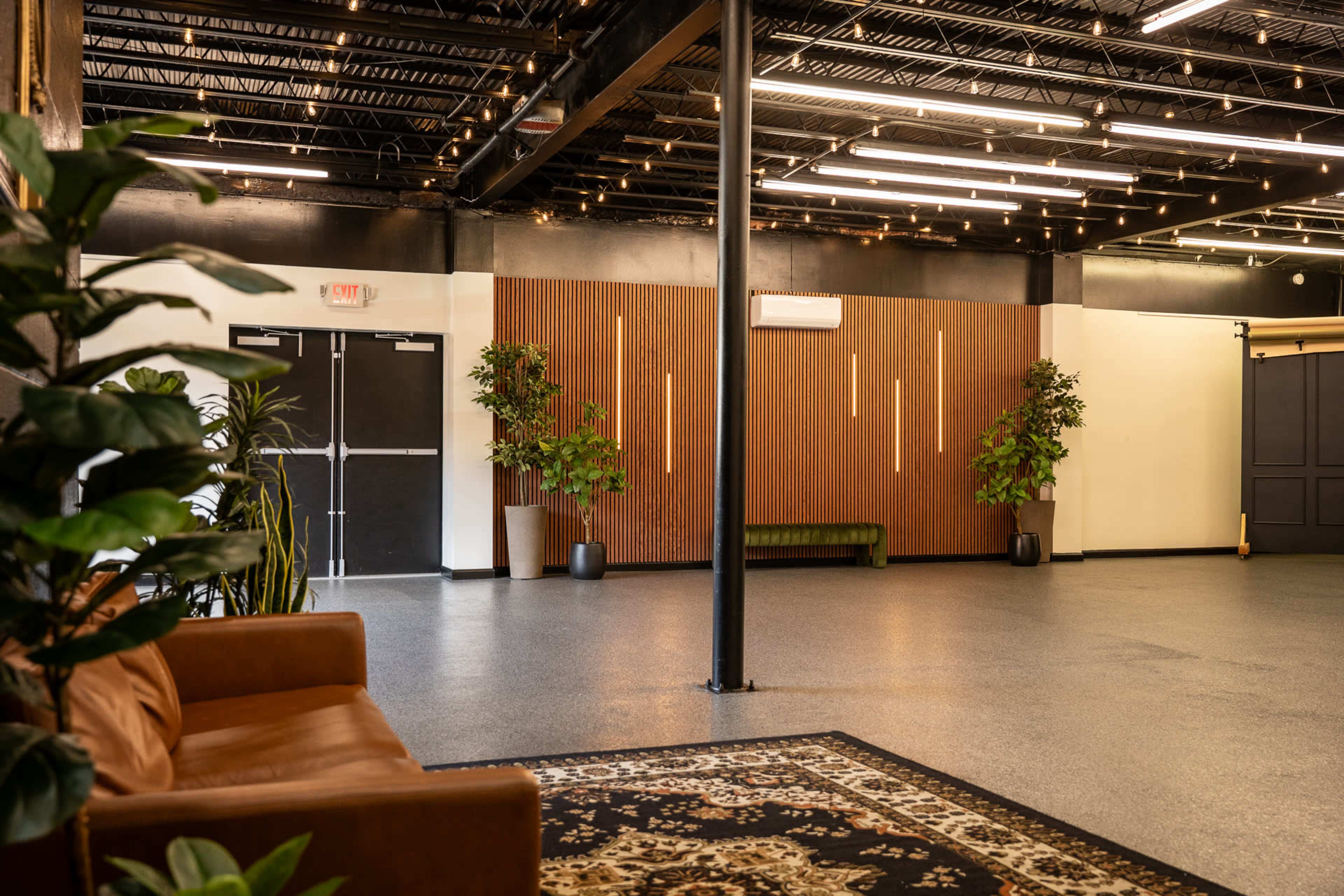 The image shows a spacious indoor venue with a wood-paneled wall, potted plants, and a brown couch under a ceiling adorned with hanging lights.