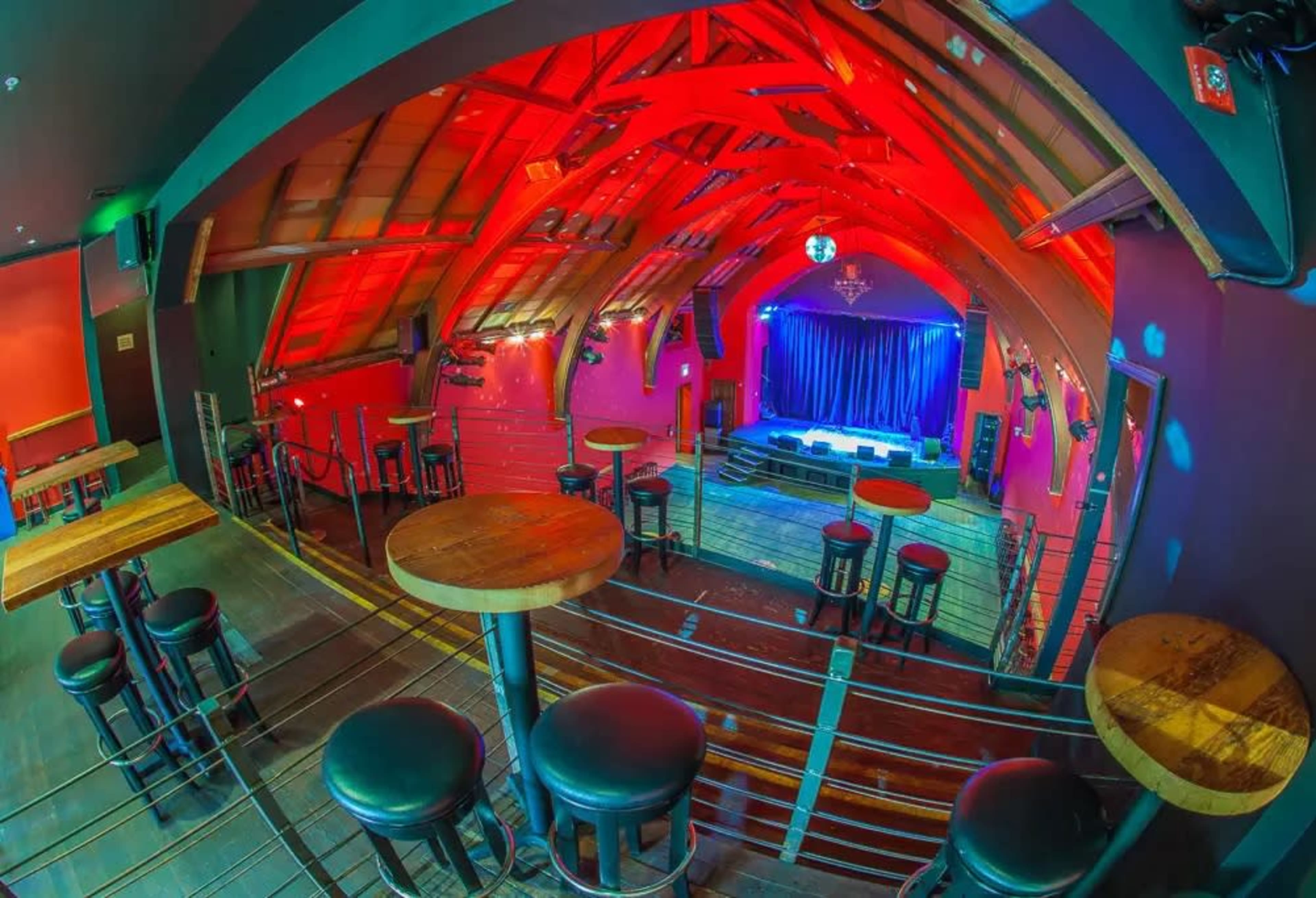 The image shows an interior view of a lively venue with a stage, bar stools, and colorful lighting.