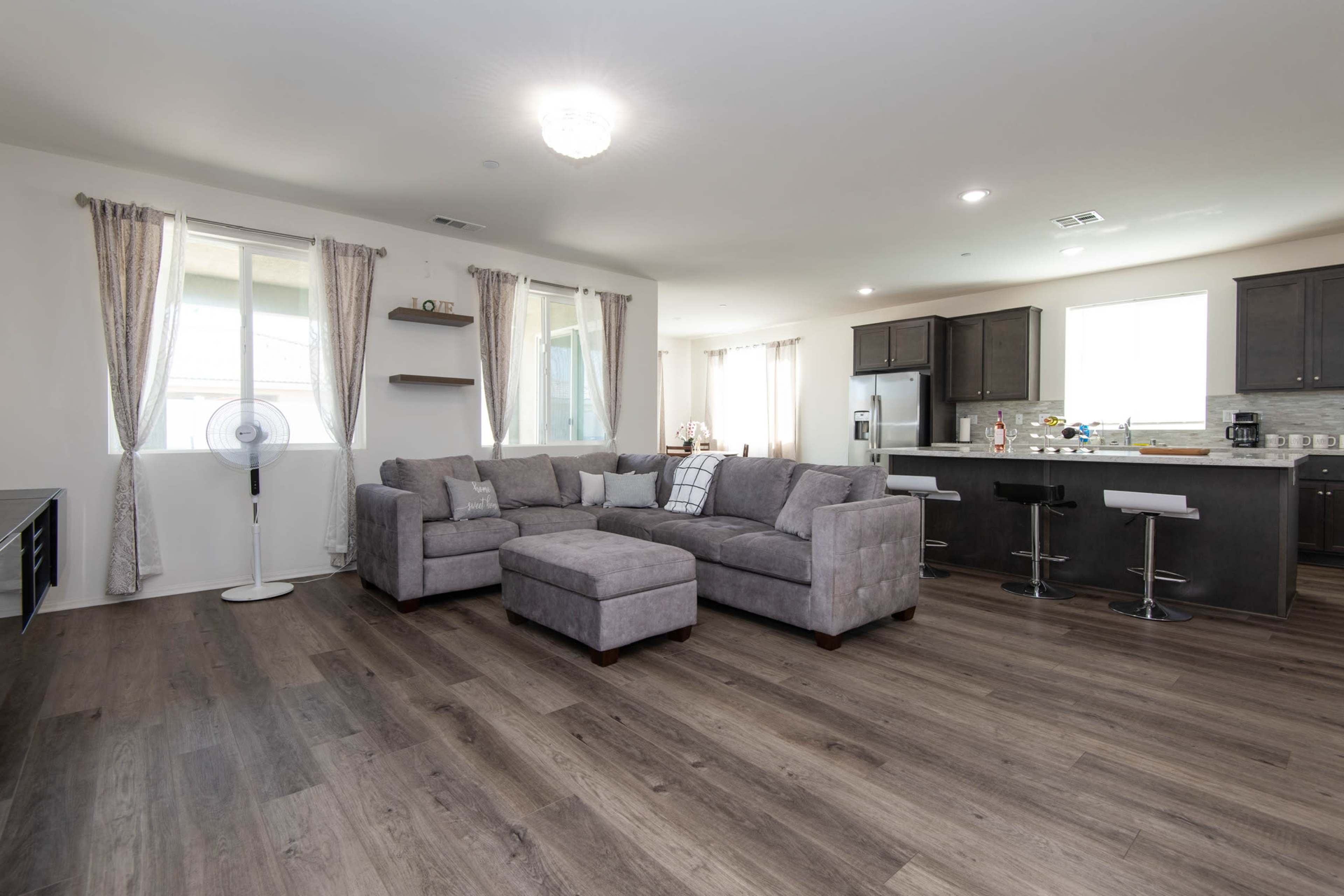 A spacious living room features a gray sectional sofa, wooden floors, and a modern kitchen with dark cabinetry and stainless steel appliances.