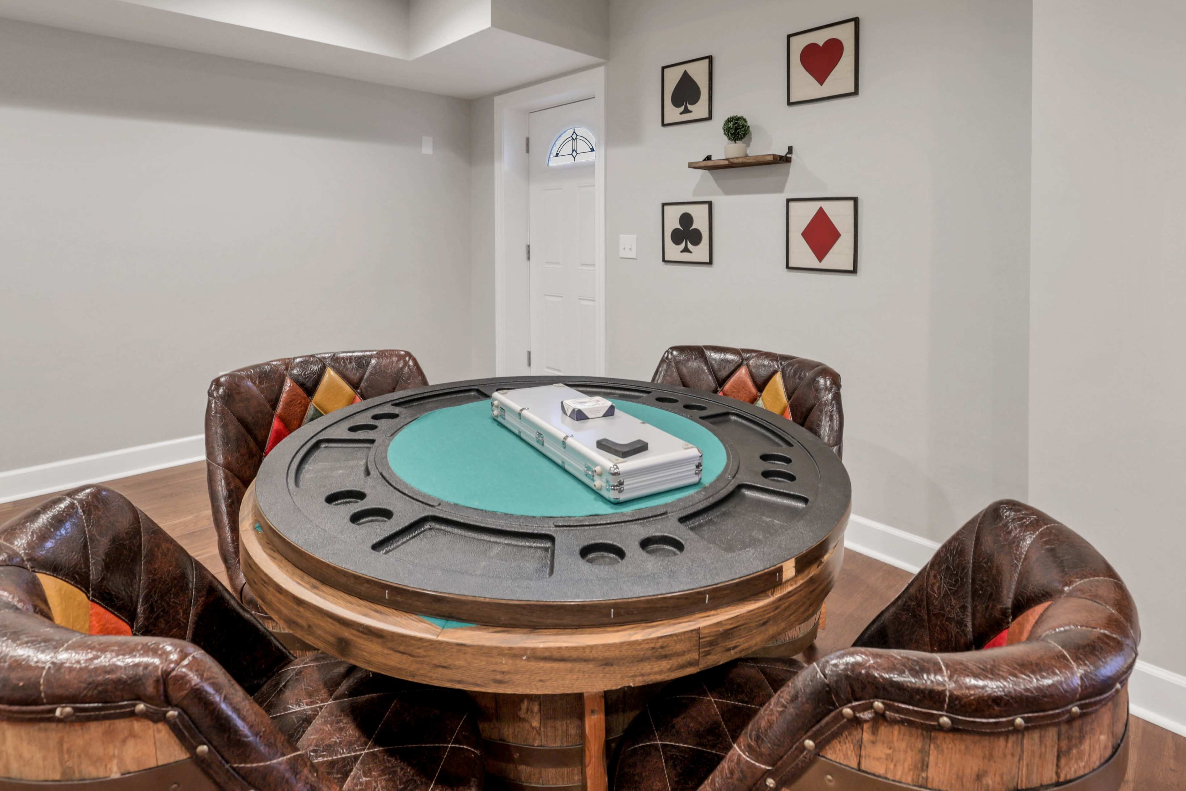 A round poker table with four upholstered chairs is set up in a well-lit room, featuring wall decorations of playing card symbols.