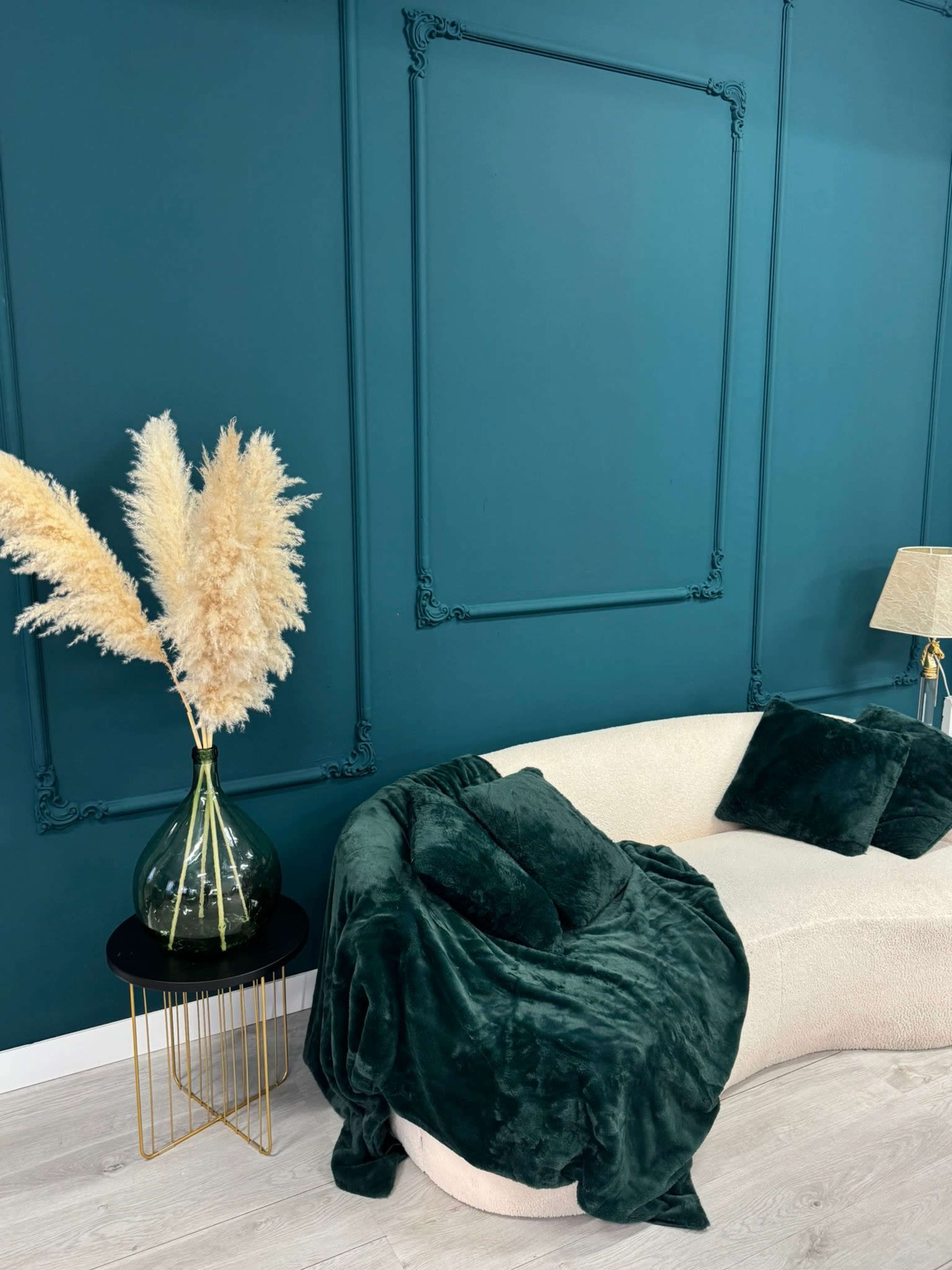 A curved cream sofa is draped with a dark green blanket, accompanied by green pillows, beside a glass vase filled with pampas grass and a lamp on a gold side table, set against a deep teal wall with ornate molding.