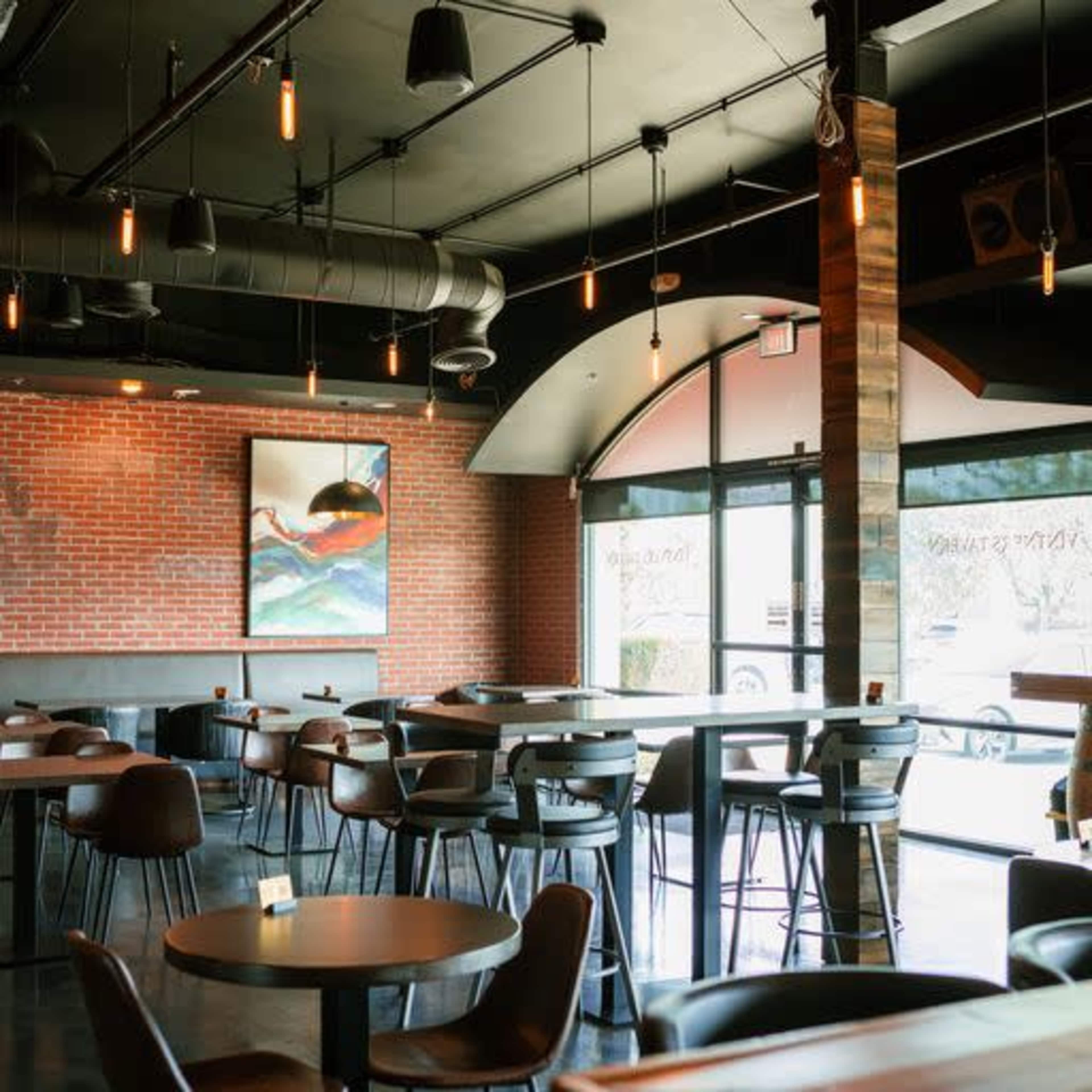 The image shows a modern restaurant interior with wooden chairs, tables, and a large window letting in natural light, featuring exposed light bulbs and a brick wall adorned with a piece of art.