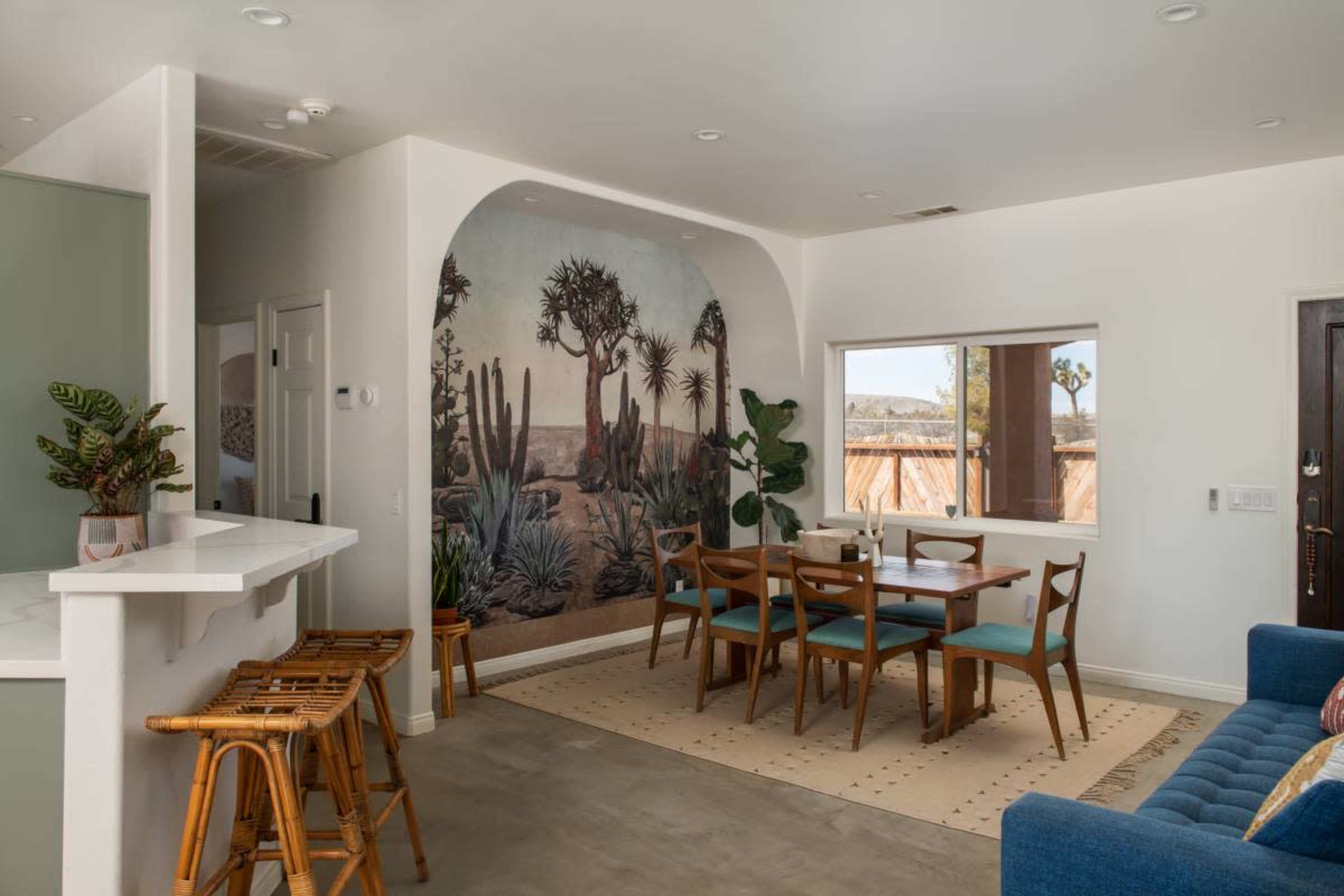 The image shows a dining area with a wooden table surrounded by blue chairs, a mural depicting cacti on one wall, and a bar counter with bamboo stools.