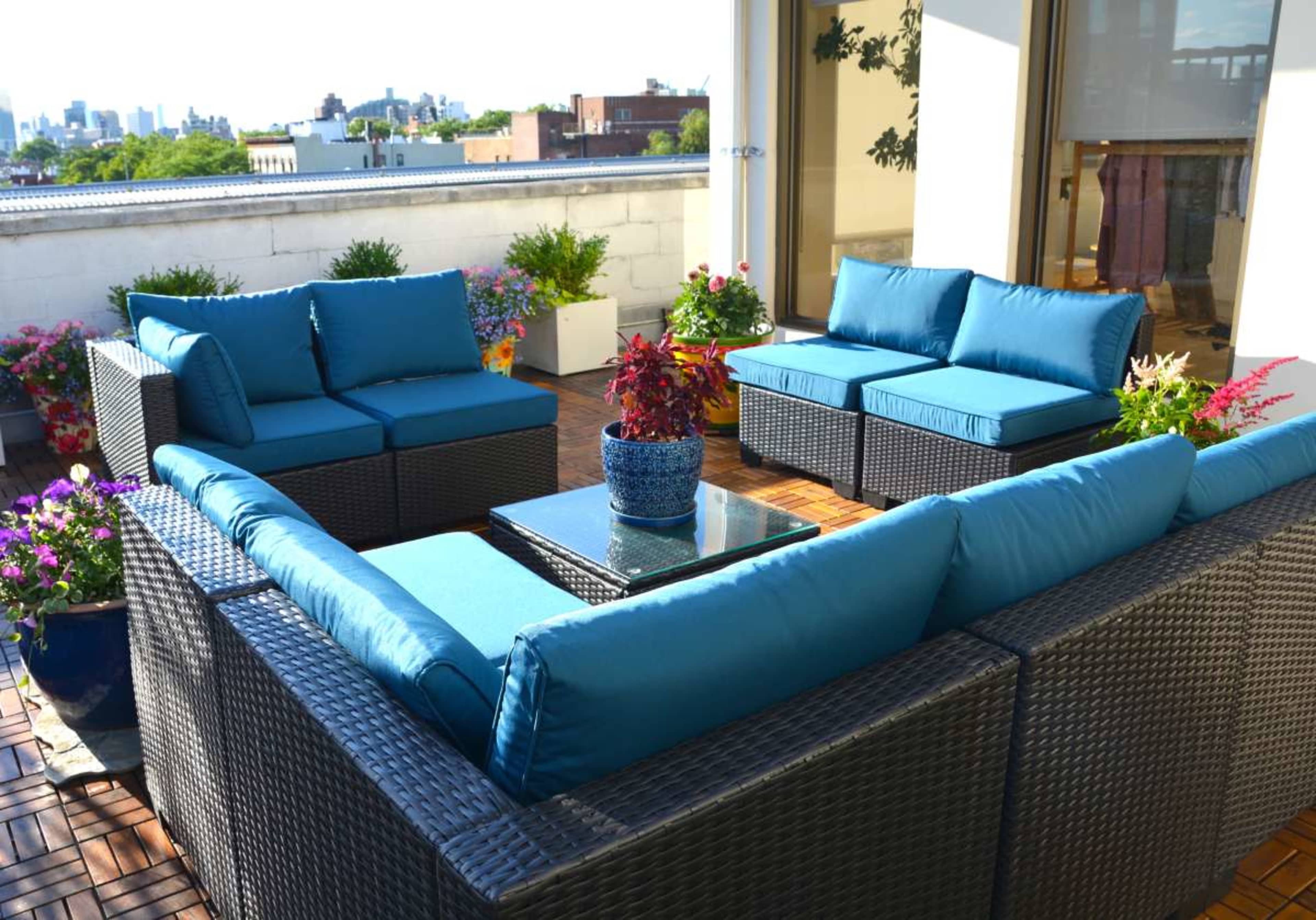 A rooftop terrace features a seating arrangement with blue cushions, surrounded by potted plants and a view of the city skyline.