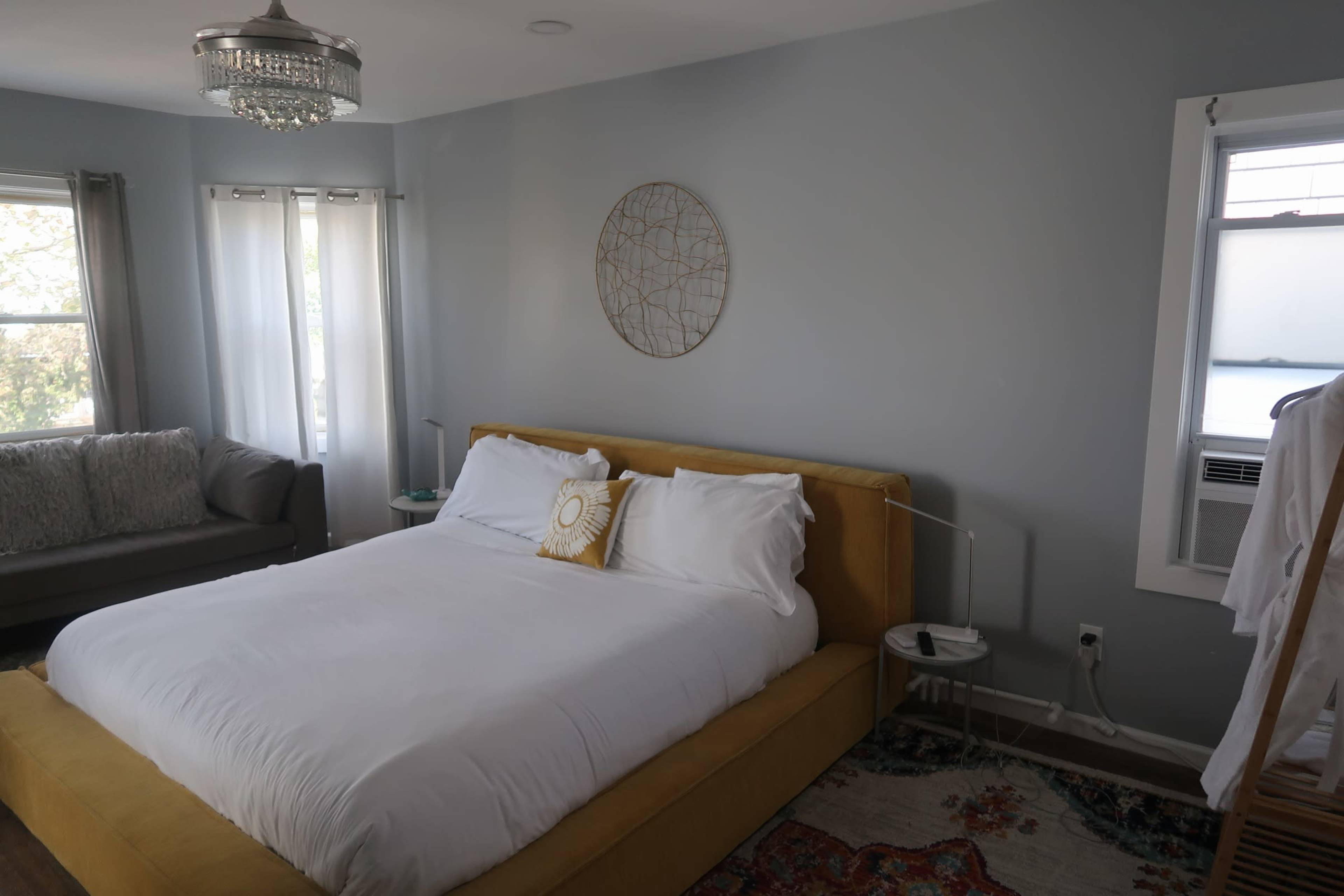A modern bedroom featuring a yellow upholstered bed with white bedding, a gray sofa, and a large round wall decoration.