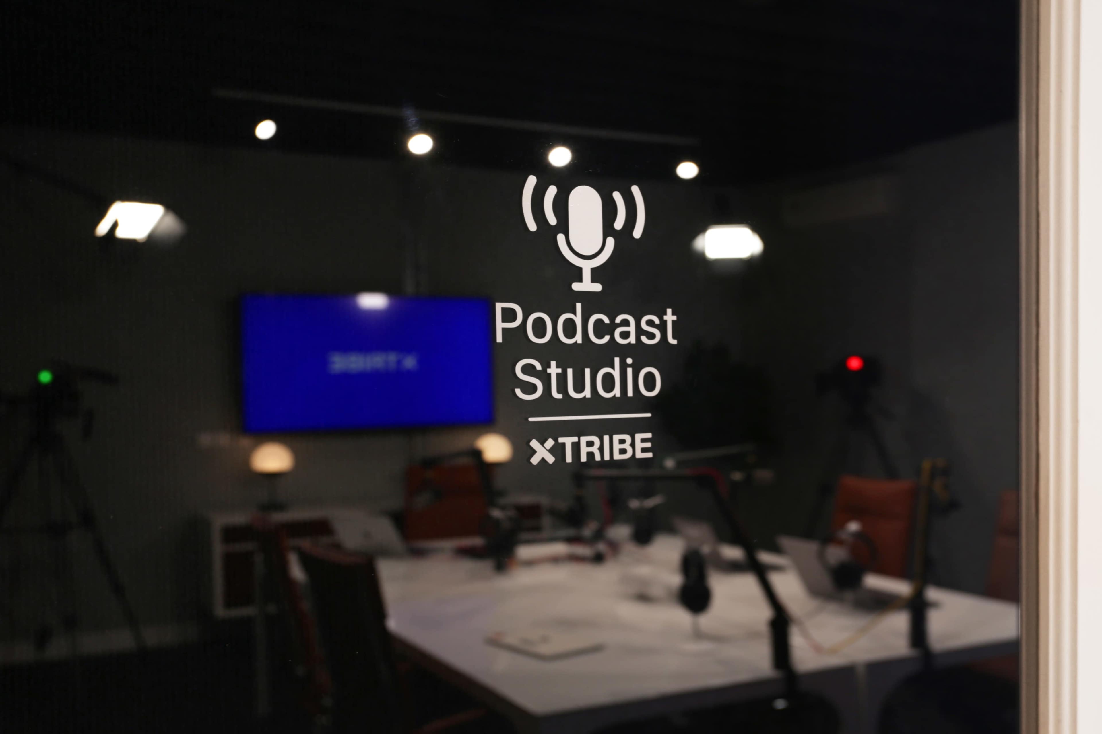 A glass door with the words "Podcast Studio" and "TRIBE" etched on it, showing a modern recording space with microphones and a large screen visible inside.