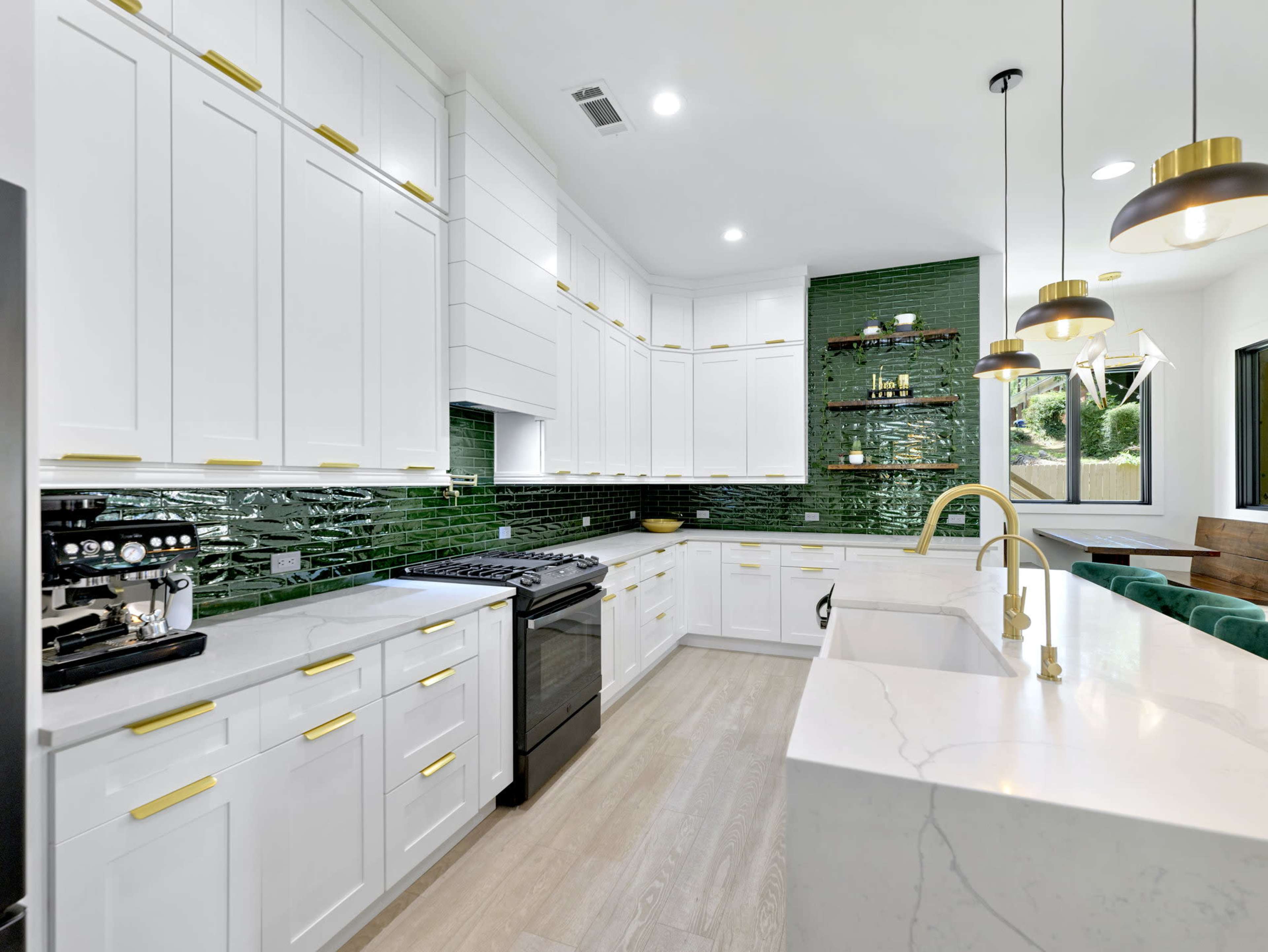 The image shows a modern kitchen featuring white cabinets with gold hardware, a green tiled backsplash, and a large island with a white countertop.