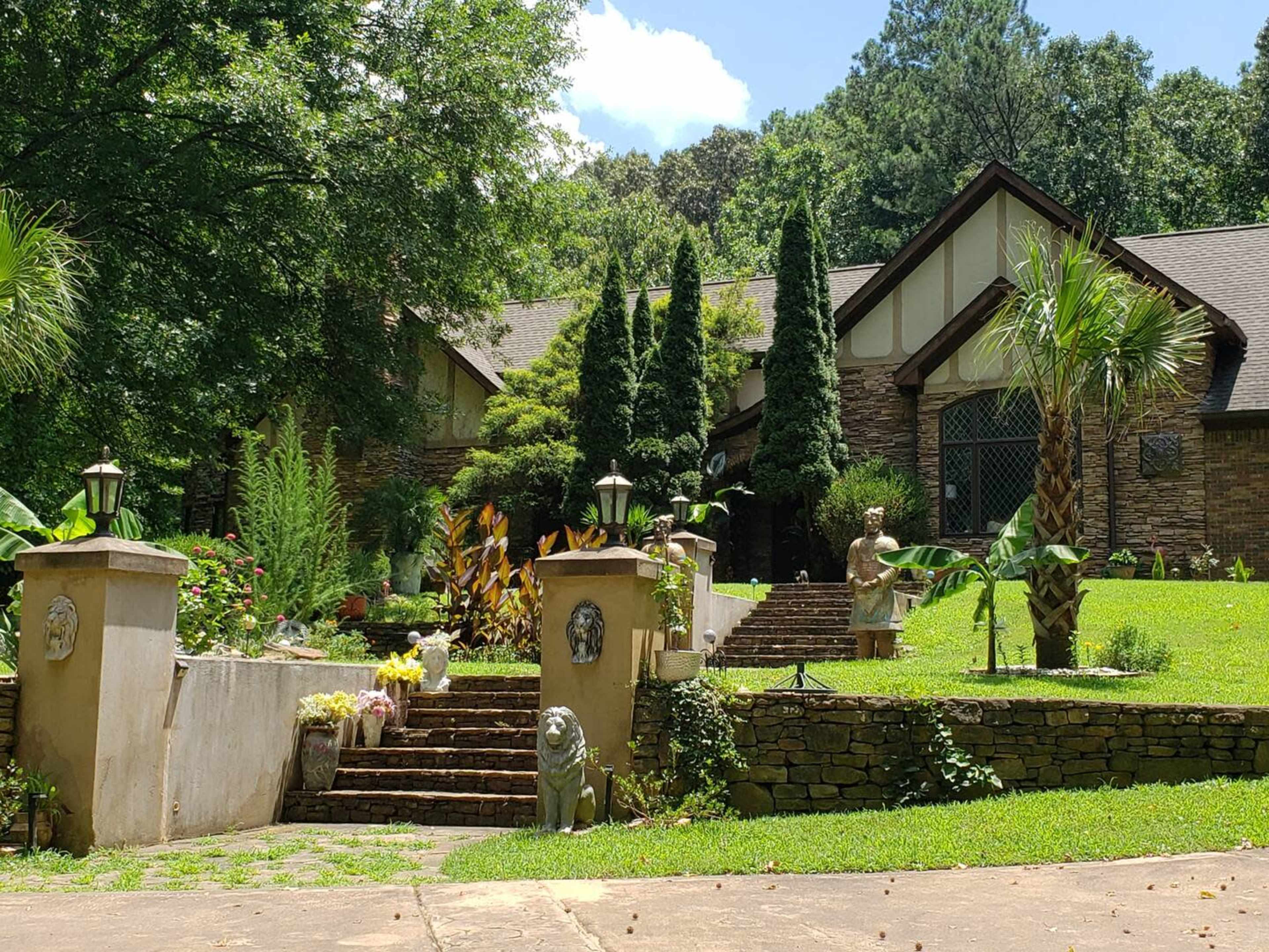 A large, stone-faced house is set atop a tiered lawn with palm trees and decorative statues lining the path to the entrance.