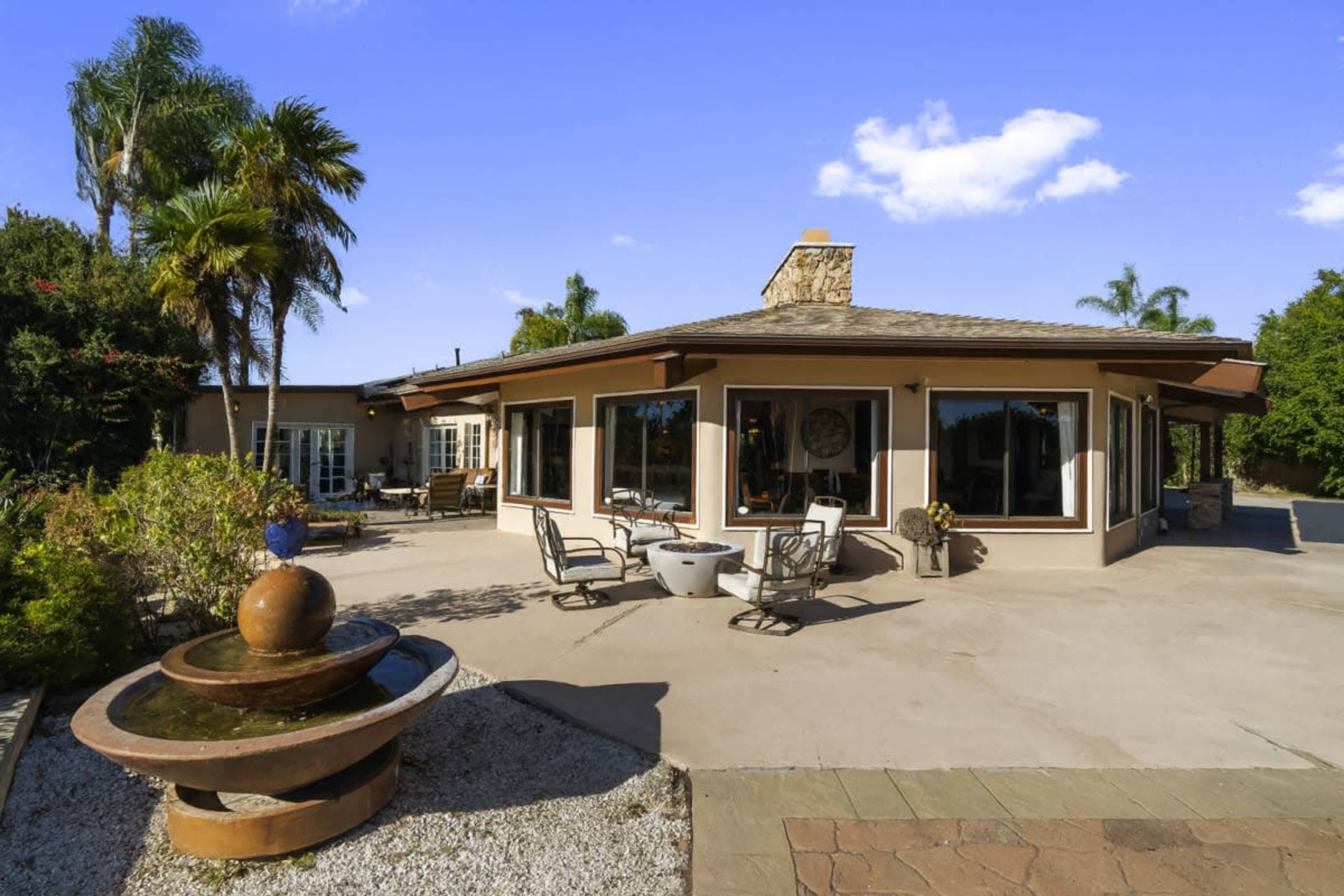 The image shows a modern single-story home with large windows and a patio area featuring a fountain and outdoor seating.