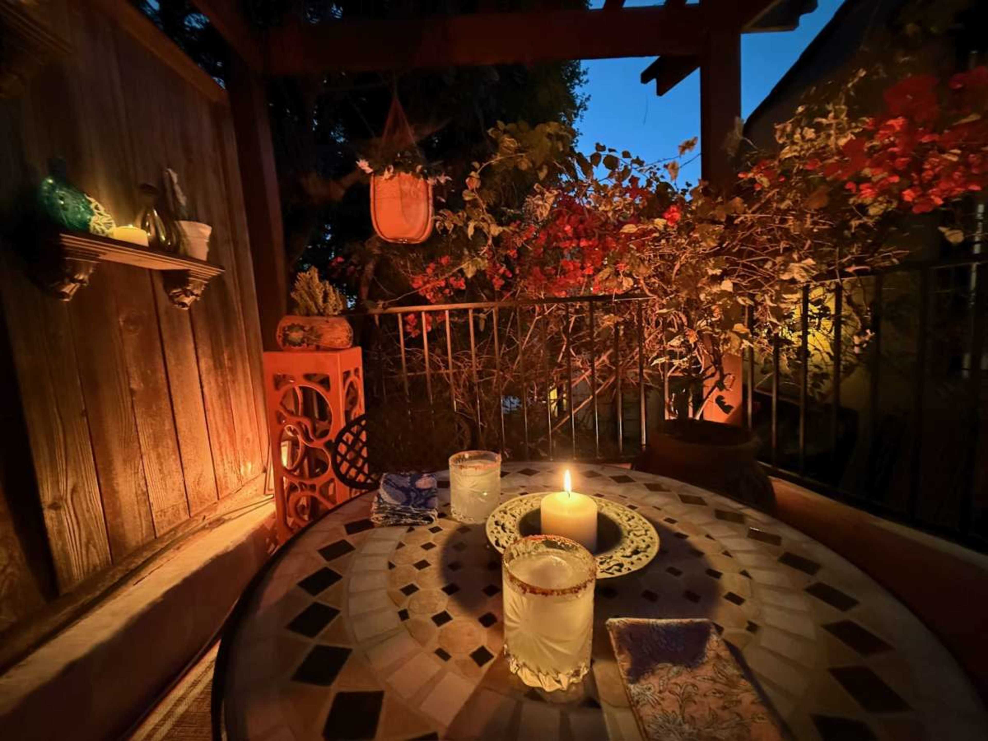 The image shows a dimly lit outdoor patio featuring a table with two glasses, a candle, and surrounding plants.