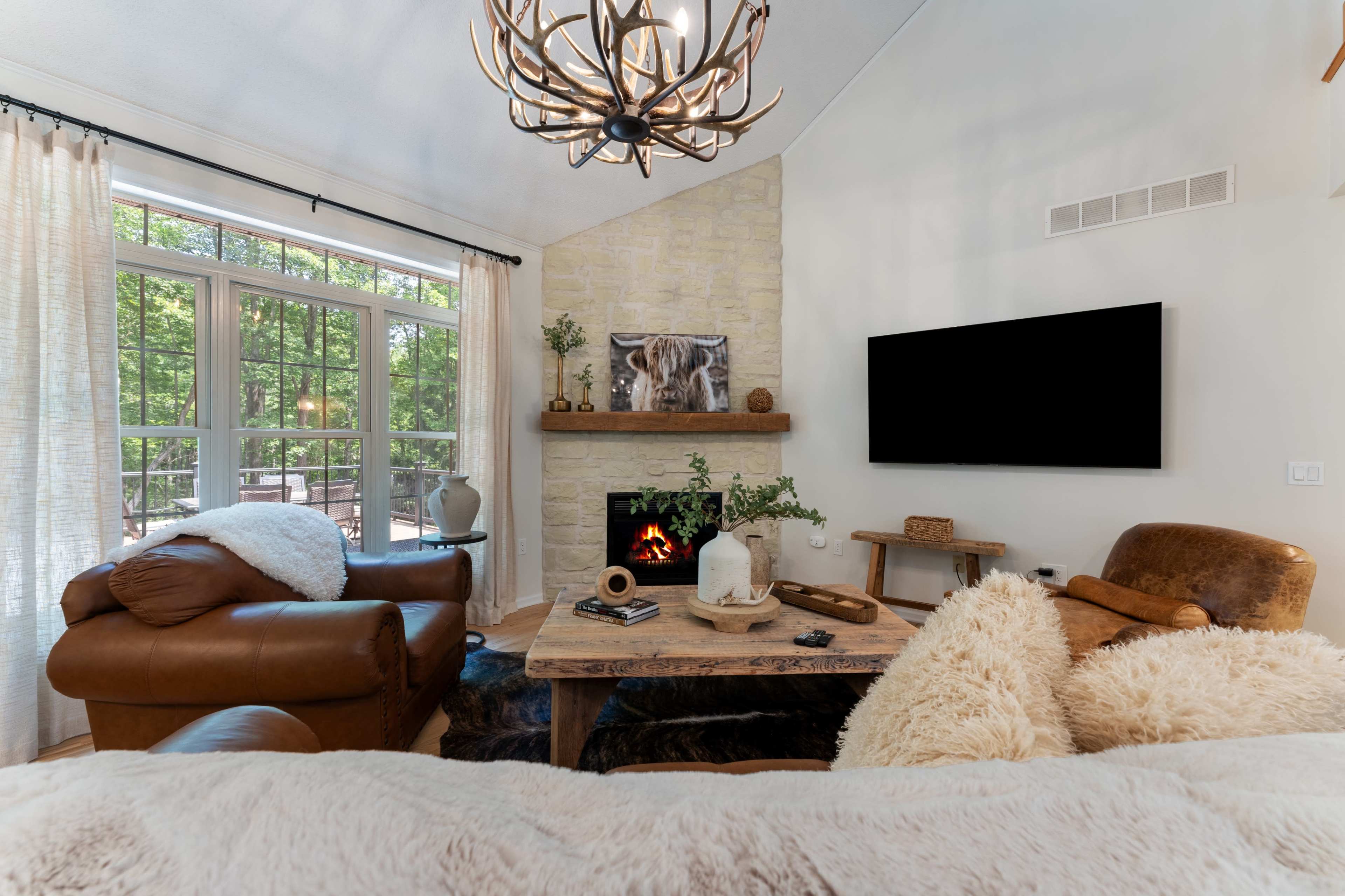 The image shows a cozy living room featuring a stone fireplace, a wooden coffee table, and two leather armchairs, with large windows allowing natural light to enter.