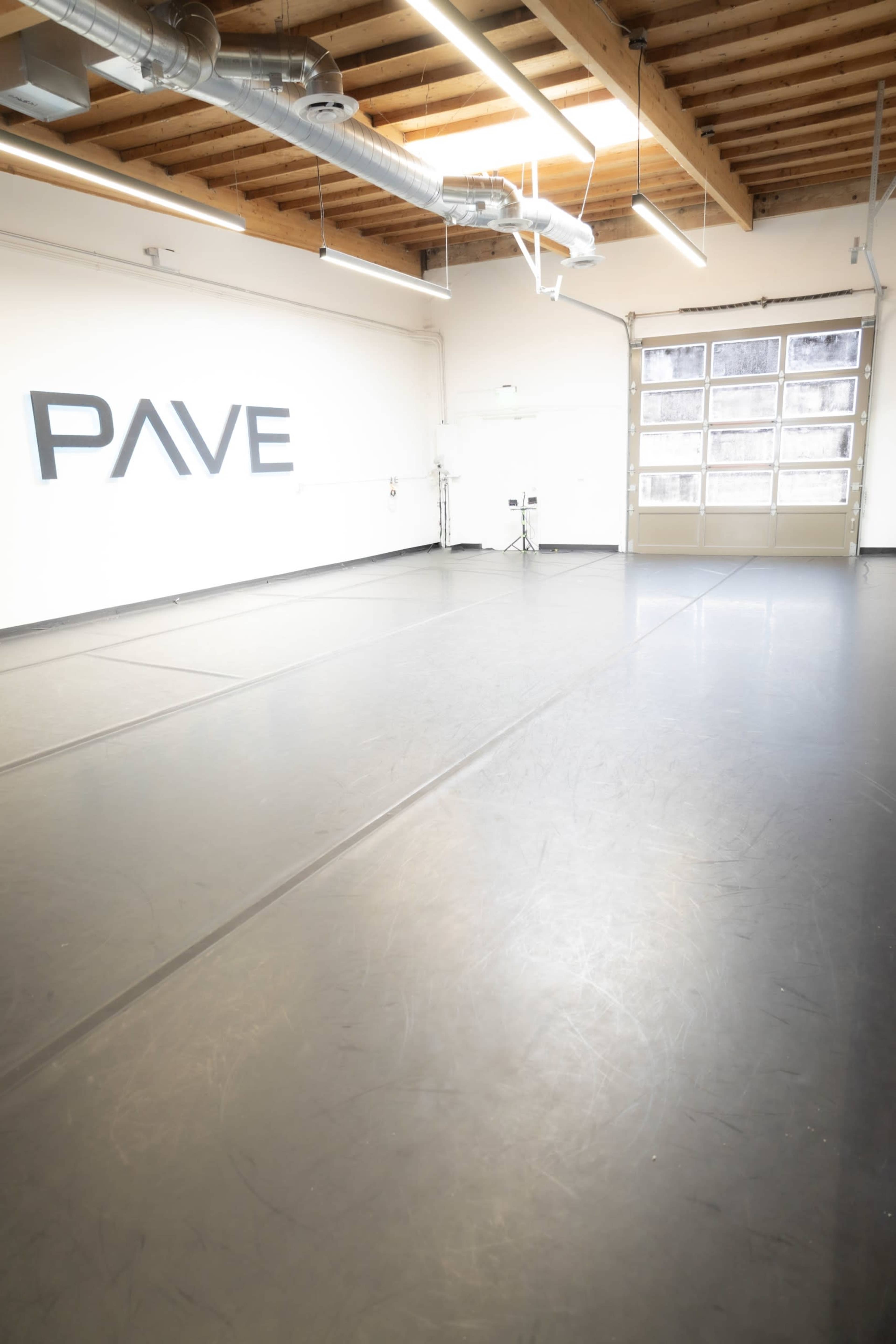 The image shows a spacious dance studio with a smooth black floor, large windows, and the word "PAVE" displayed on the wall.