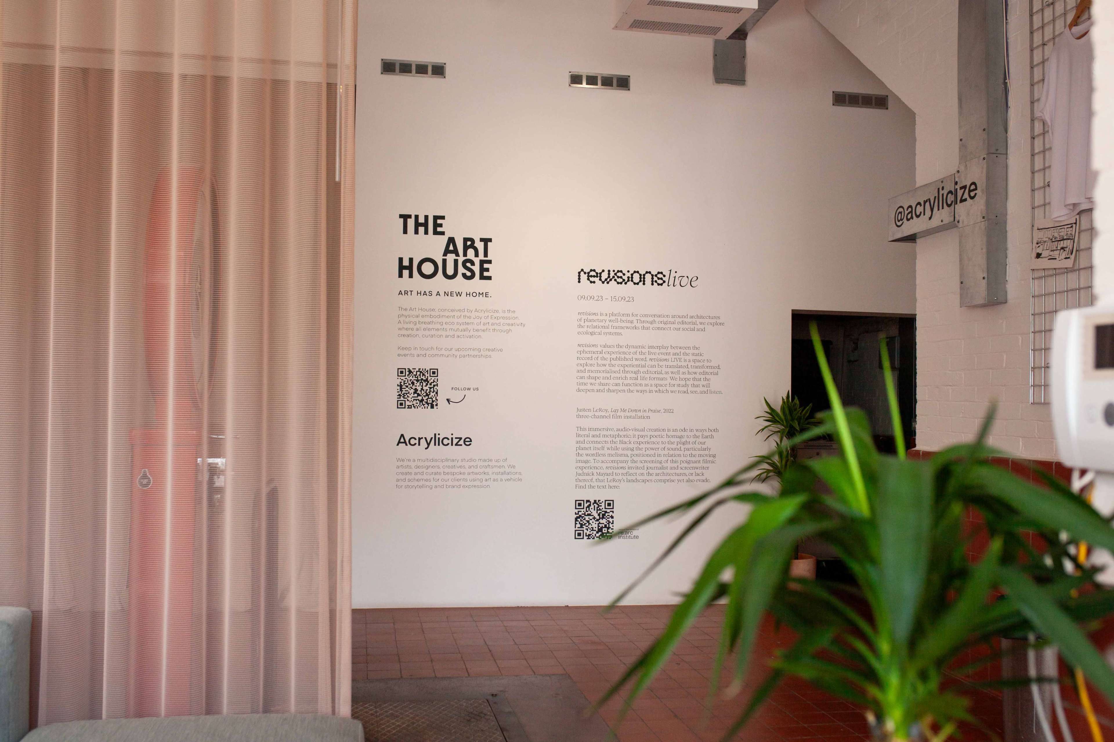 An interior space with a wall displaying informational text about "The Art House" and "Acrylcize," accompanied by QR codes, and a green plant in the foreground.