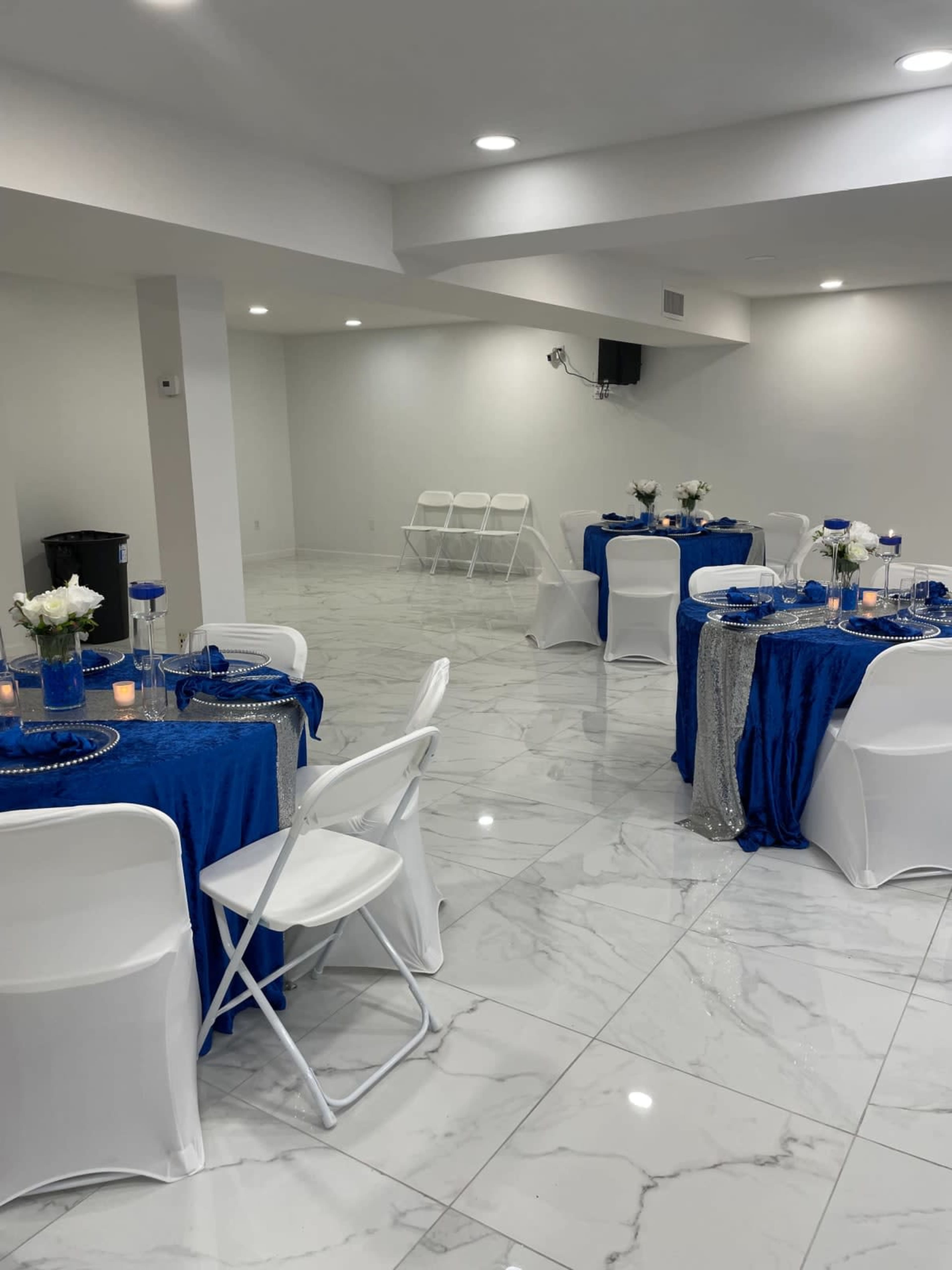 The image shows a well-lit event space with two tables covered in blue tablecloths, white chairs, and floral centerpieces.