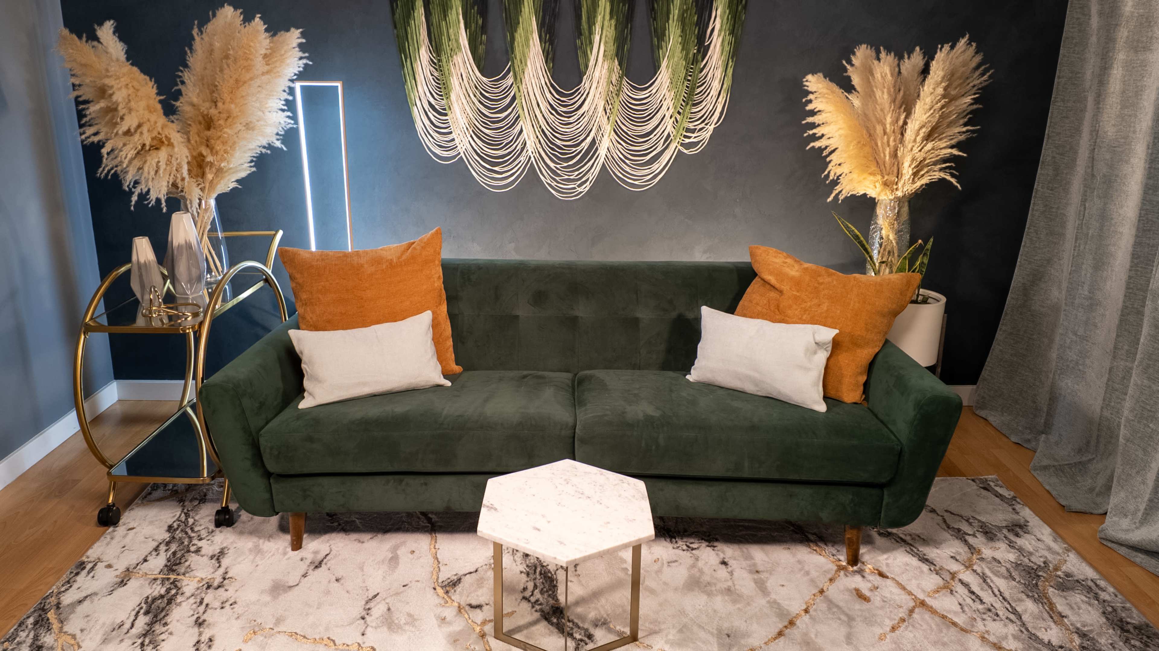 A green sofa with orange and white pillows is positioned on a marble-patterned rug in a stylish living room featuring decorative pampas grass and a gold accent table.