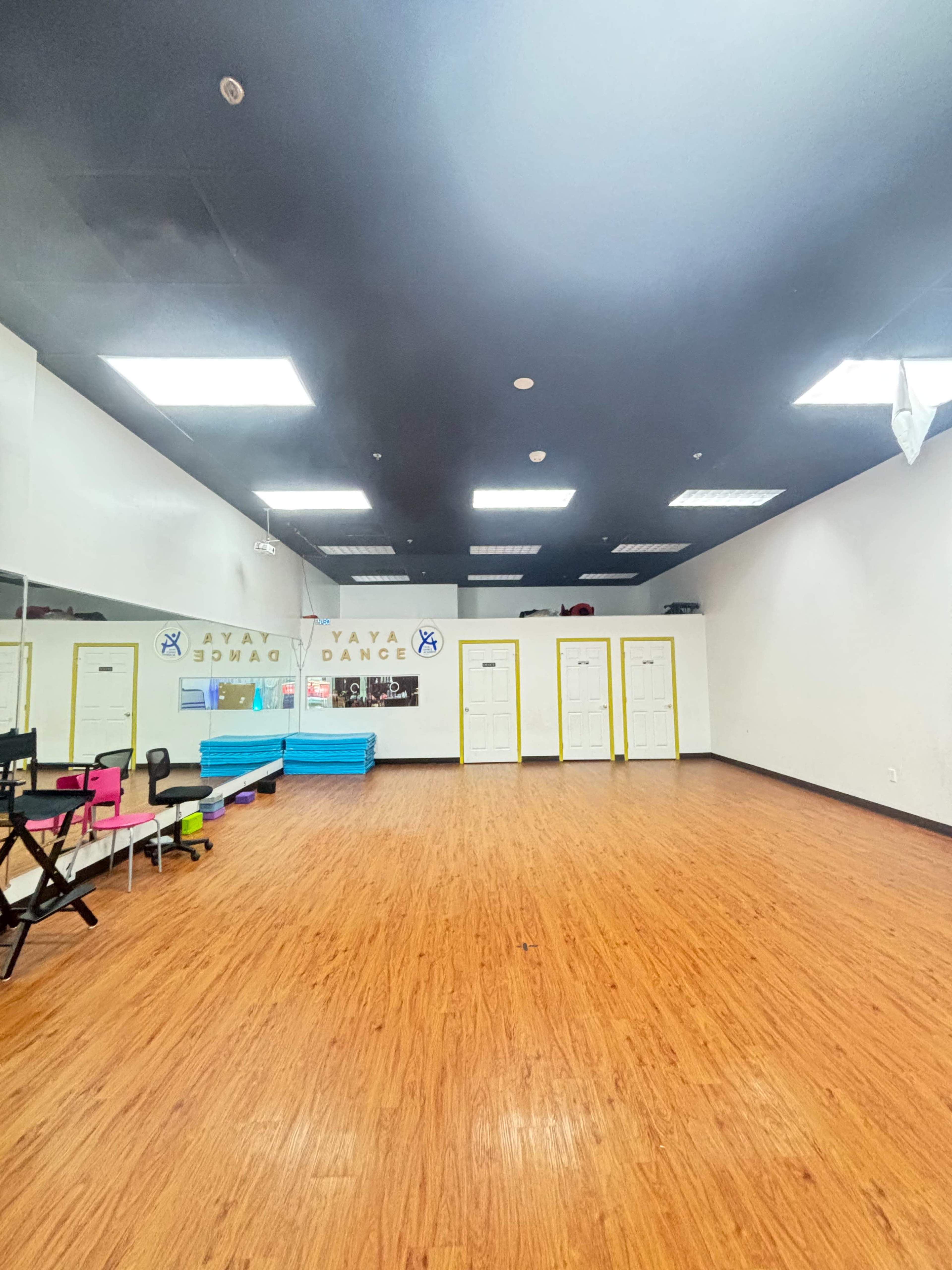 The image shows a dance studio with wooden flooring, mirrored walls, and storage areas along the back, illuminated by ceiling lights.