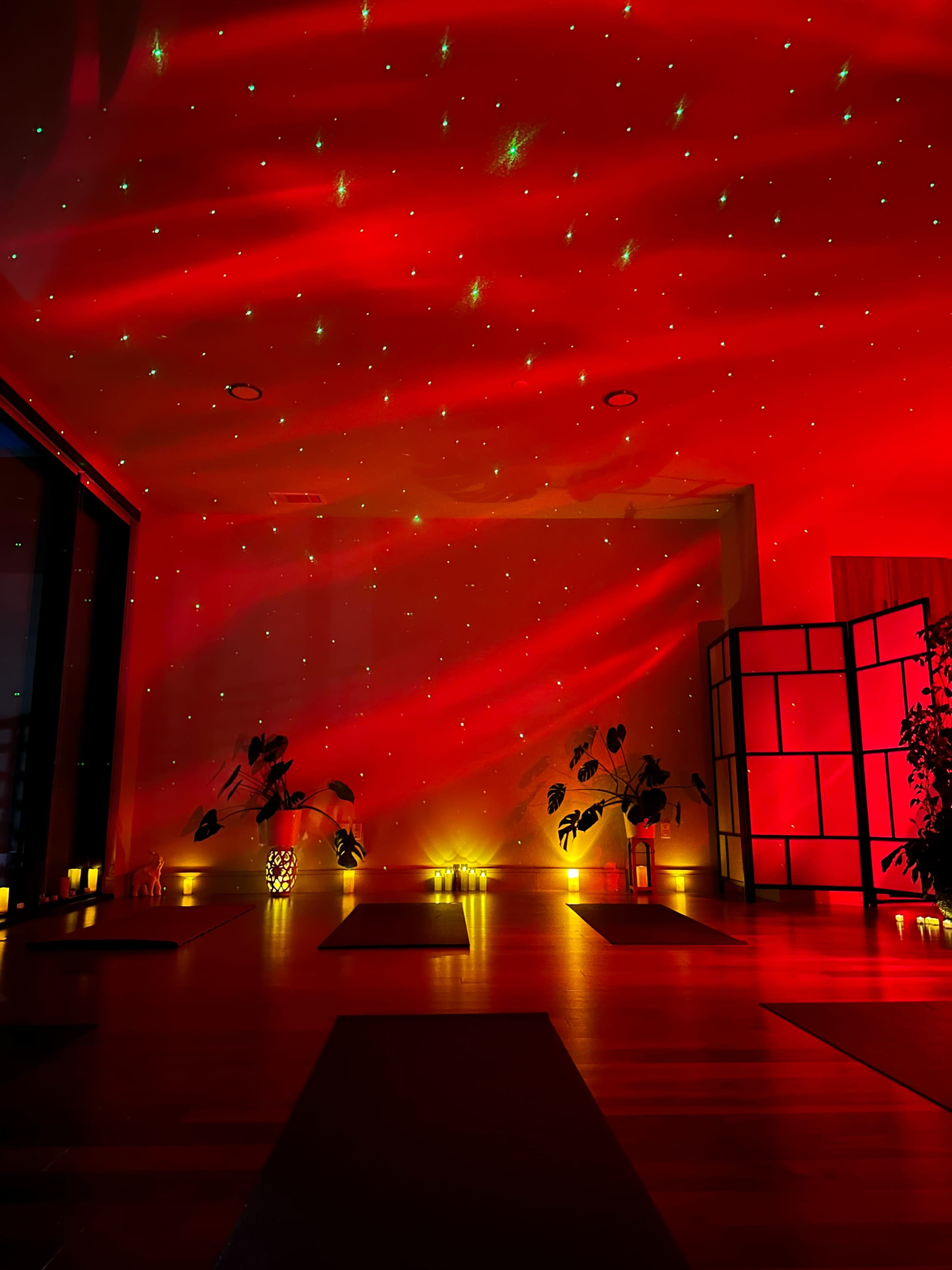 The image shows a dimly lit yoga studio with red lighting, star-like projections on the walls, and plants placed around the space.