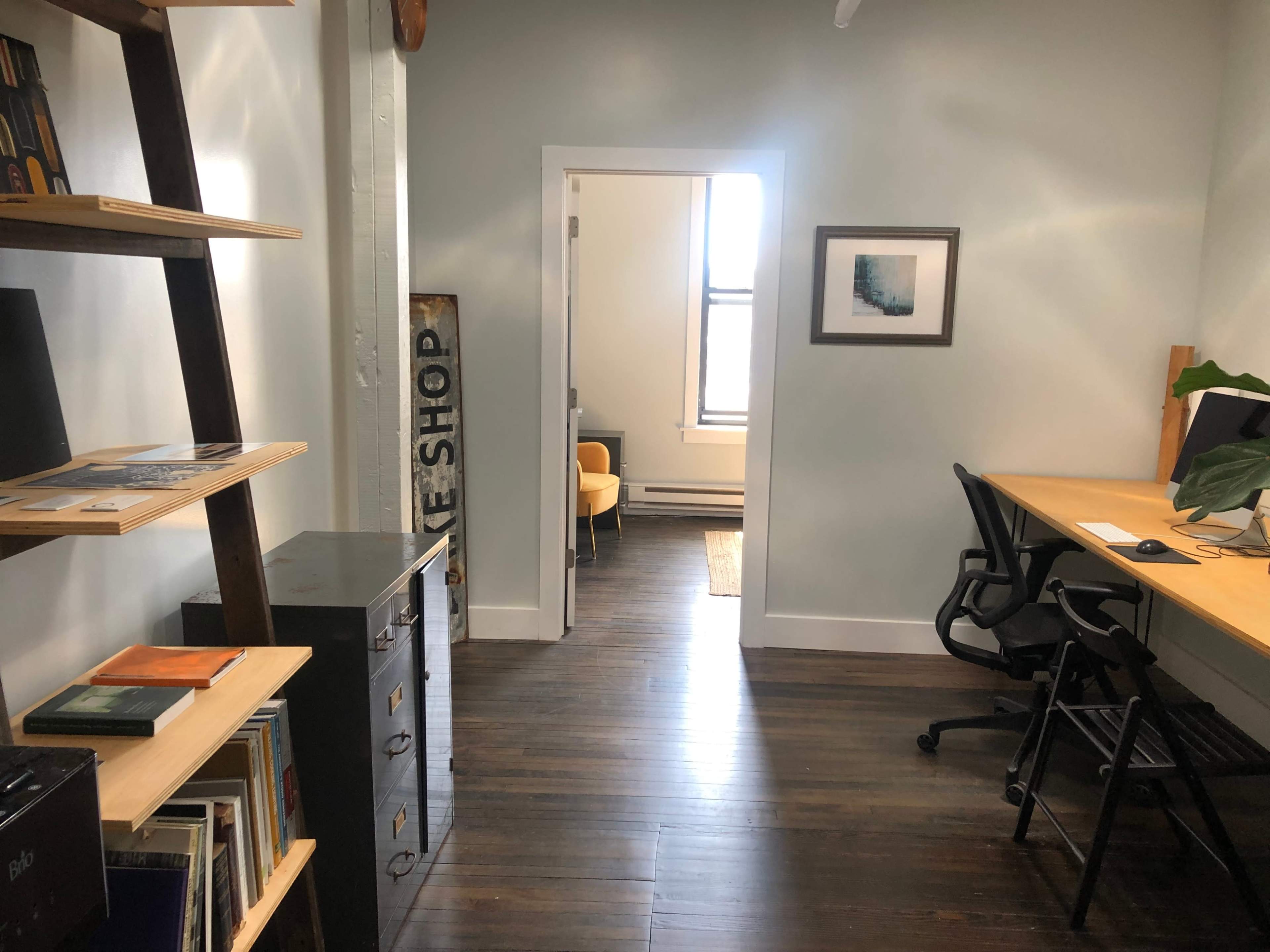 The image shows a hallway leading to a room with a desk and chairs, featuring a bookshelf and a sign on the wall.