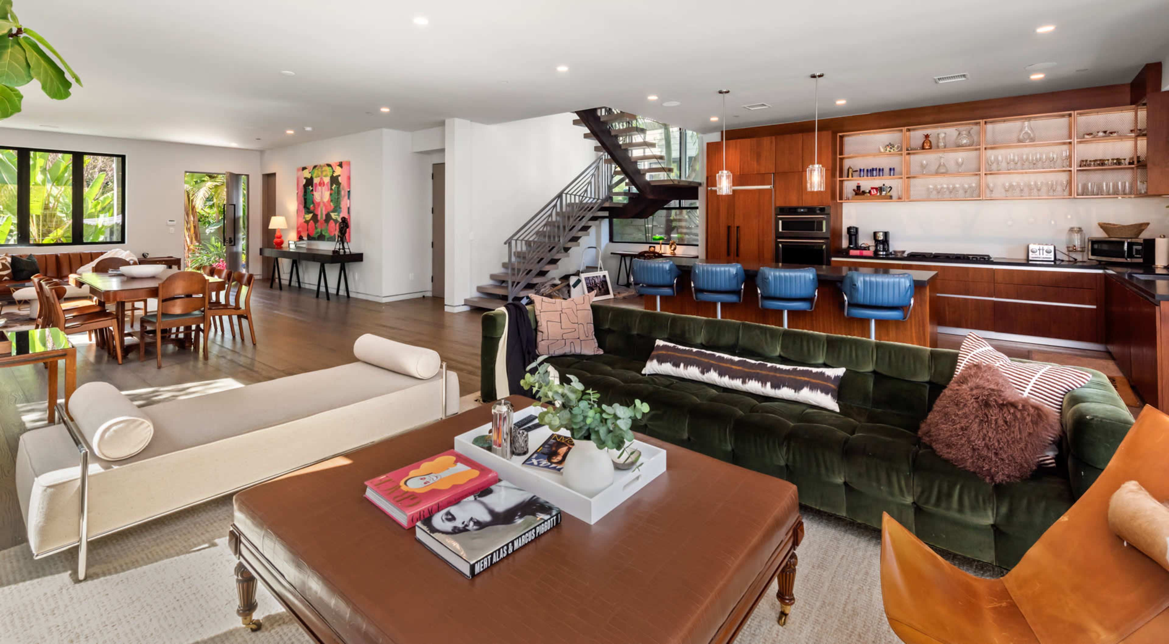 The image shows a modern open-concept living space featuring a living area with a green velvet sofa, coffee table, and a dining area with wooden chairs, all illuminated by natural light streaming through large windows.