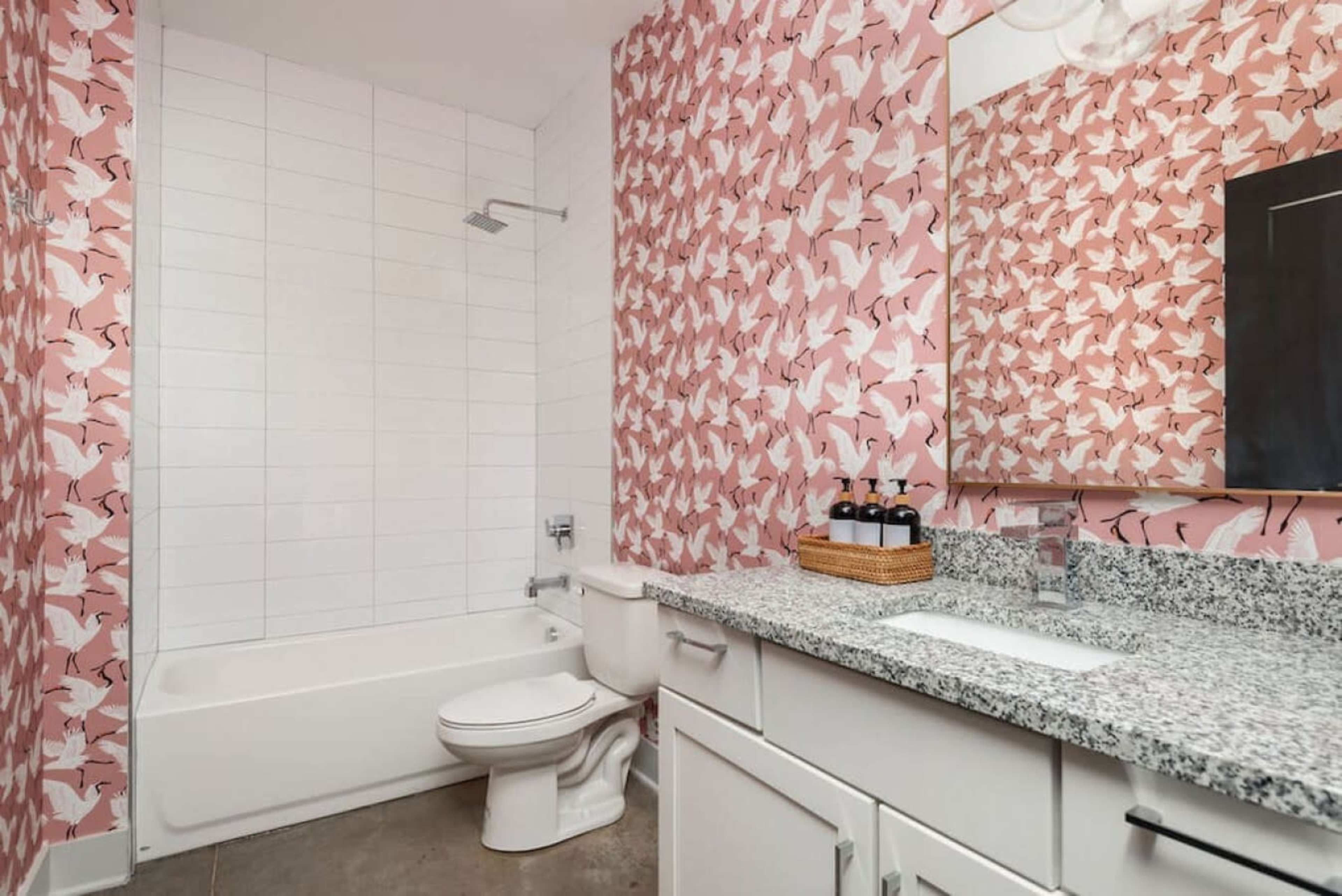 The bathroom features a bathtub with a shower, a toilet, and a vanity with a granite countertop, all set against a pink patterned wallpaper.