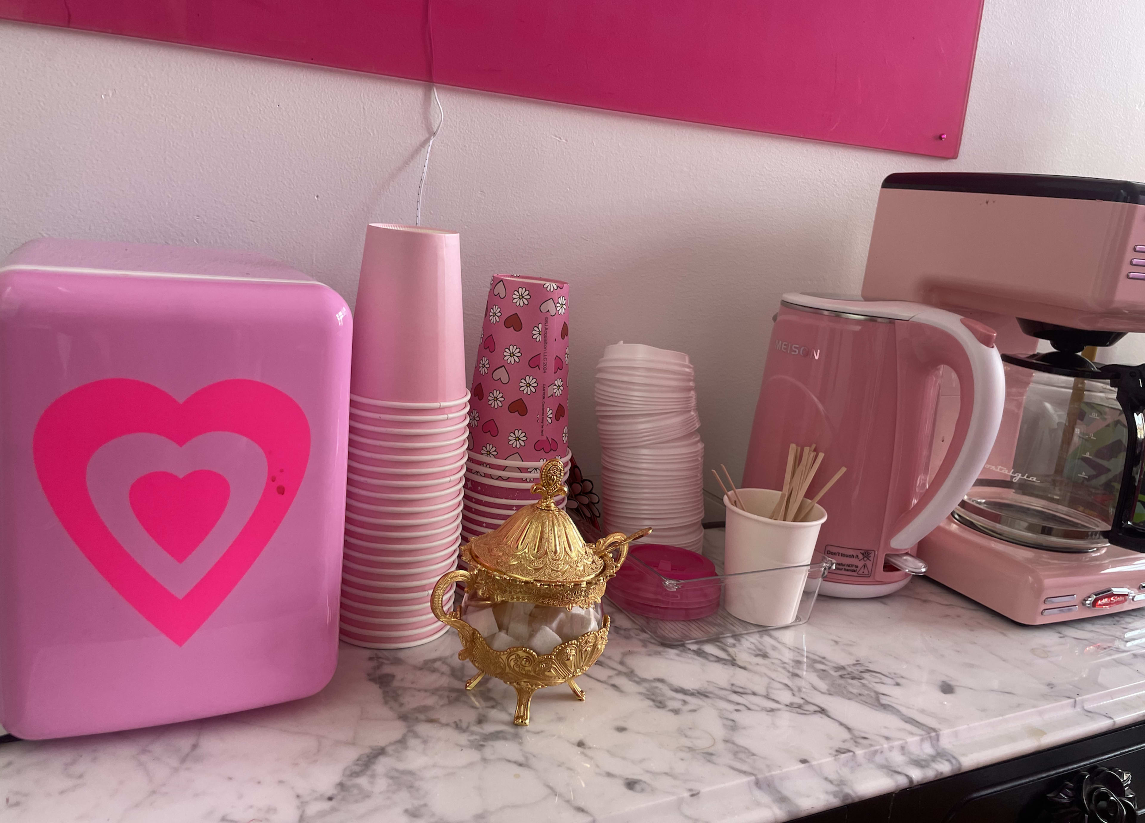 The image shows a pink kitchen countertop featuring a heart-patterned mini fridge, pink cups, a kettle, a coffee maker, and a decorative golden tea set.