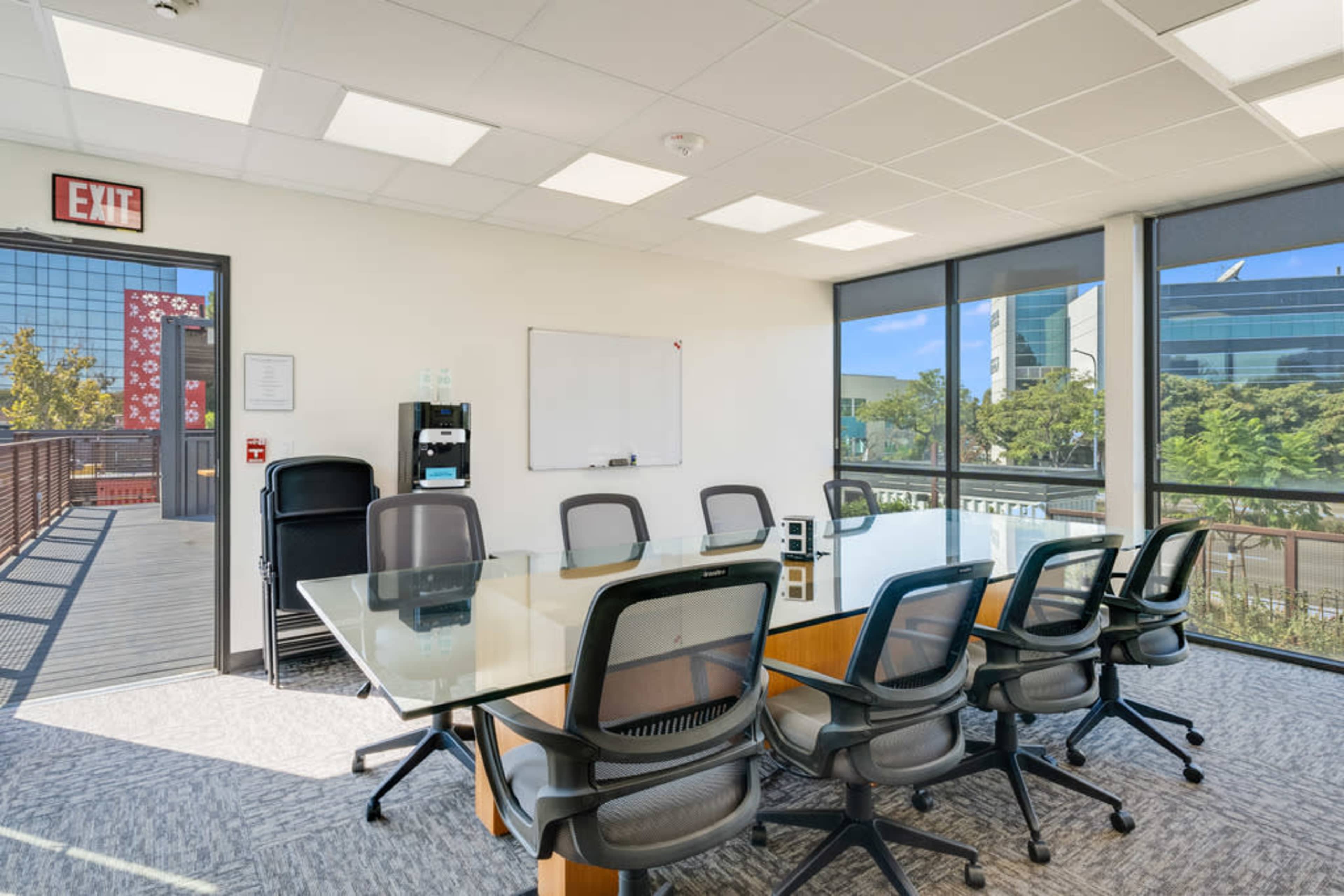 A bright conference room features a large glass table surrounded by black ergonomic chairs, with windows showcasing a view of the outdoors.
