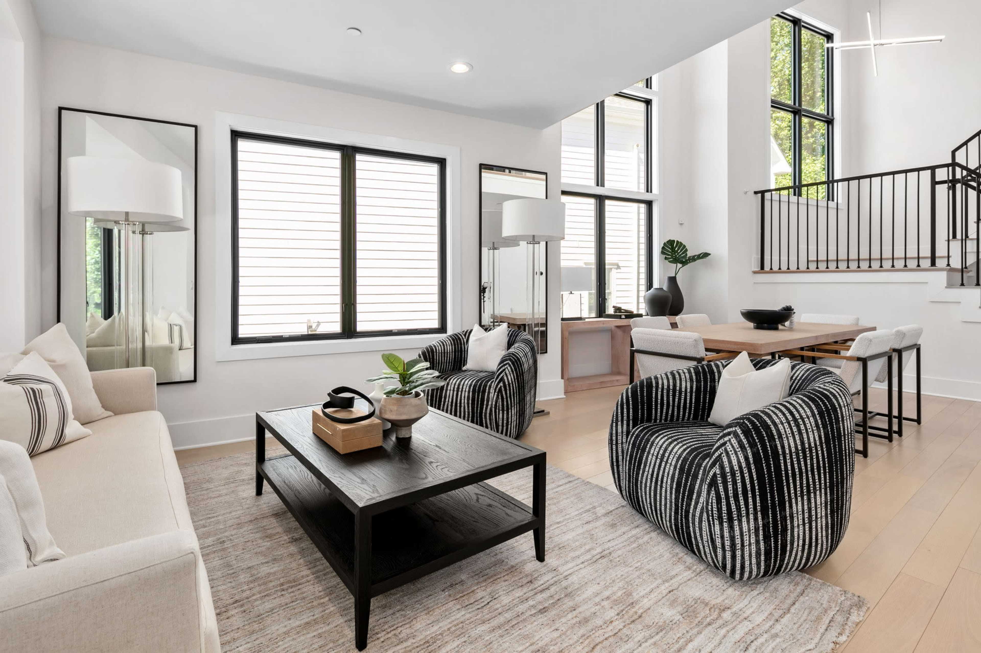 A modern living room features a beige sofa, striped armchairs, a dark coffee table, and large windows allowing natural light to enter.