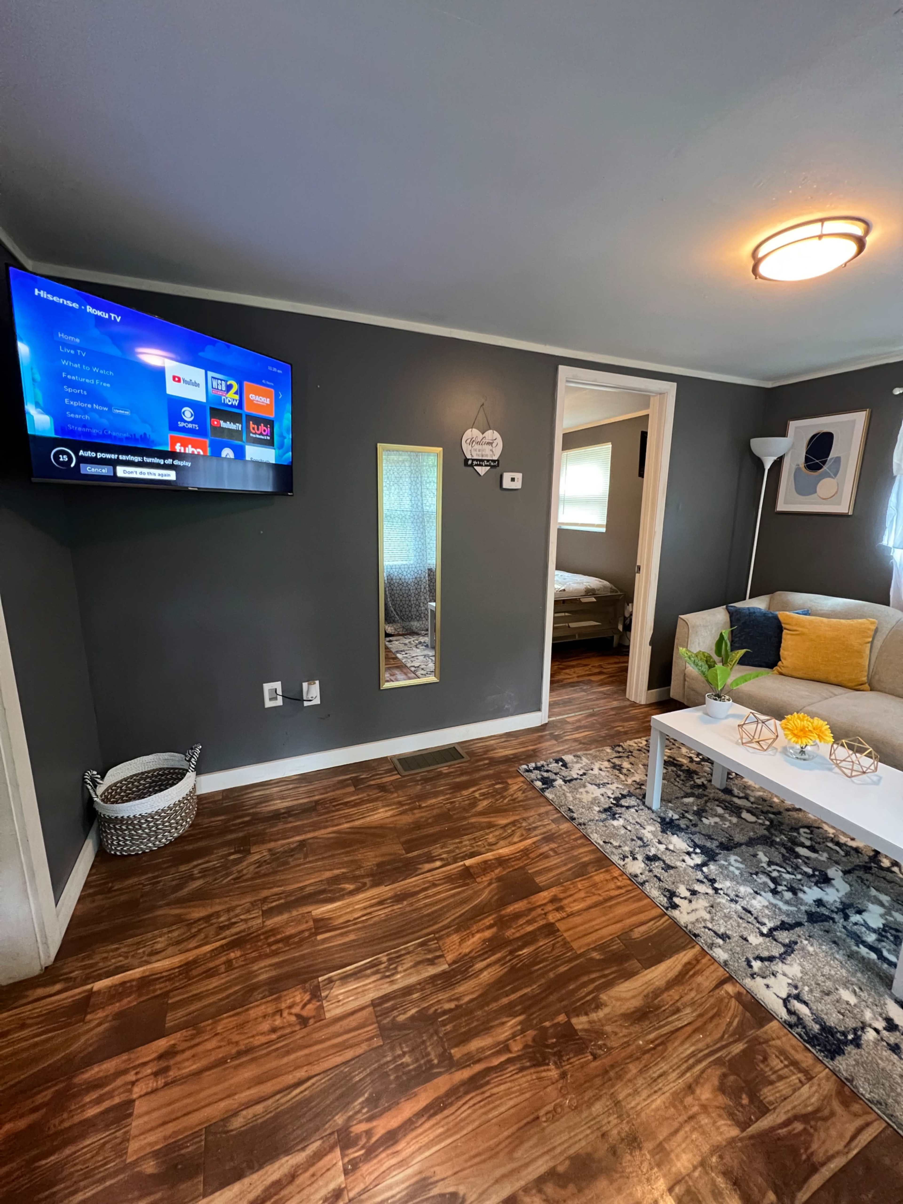 A living room with a wall-mounted TV, a mirror, a coffee table on a rug, and a sofa with yellow cushions.