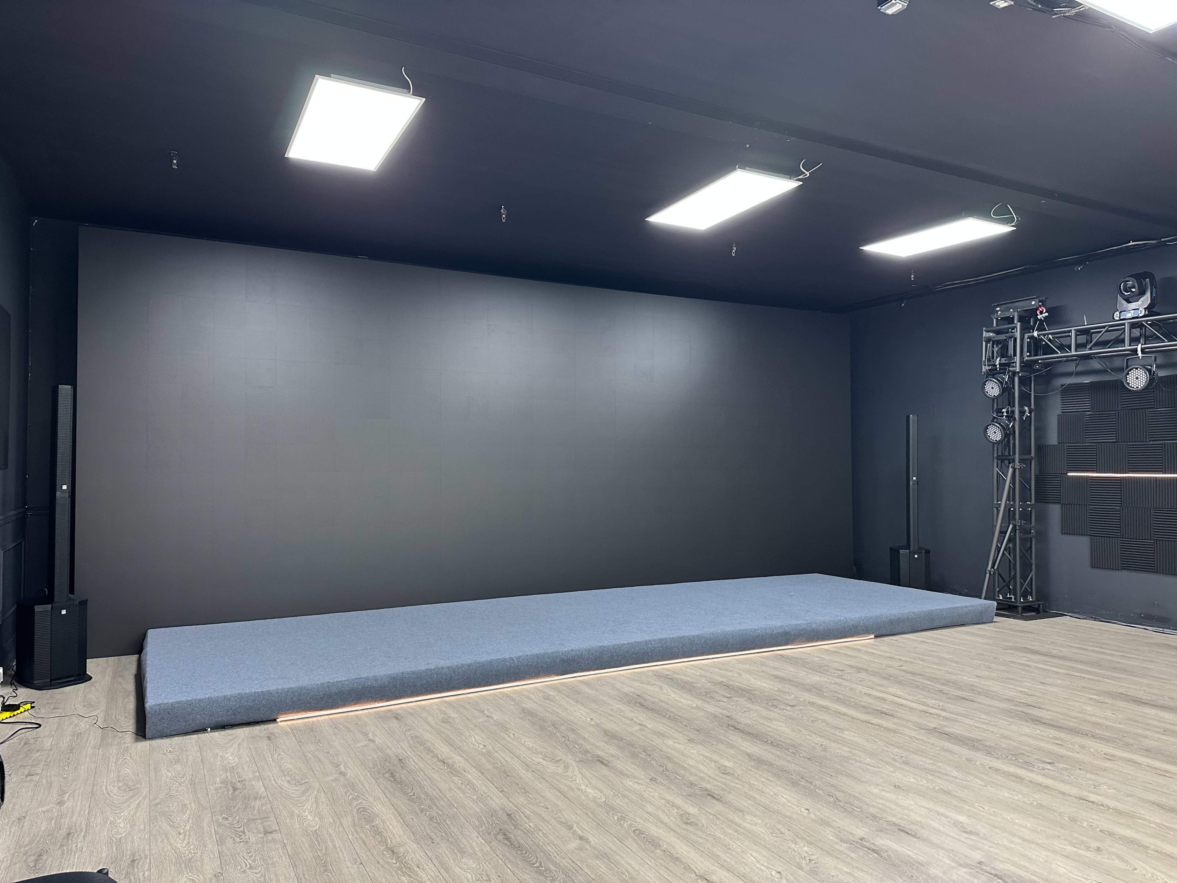 The image shows an empty studio space with a large, grey platform against a dark wall and overhead lighting.