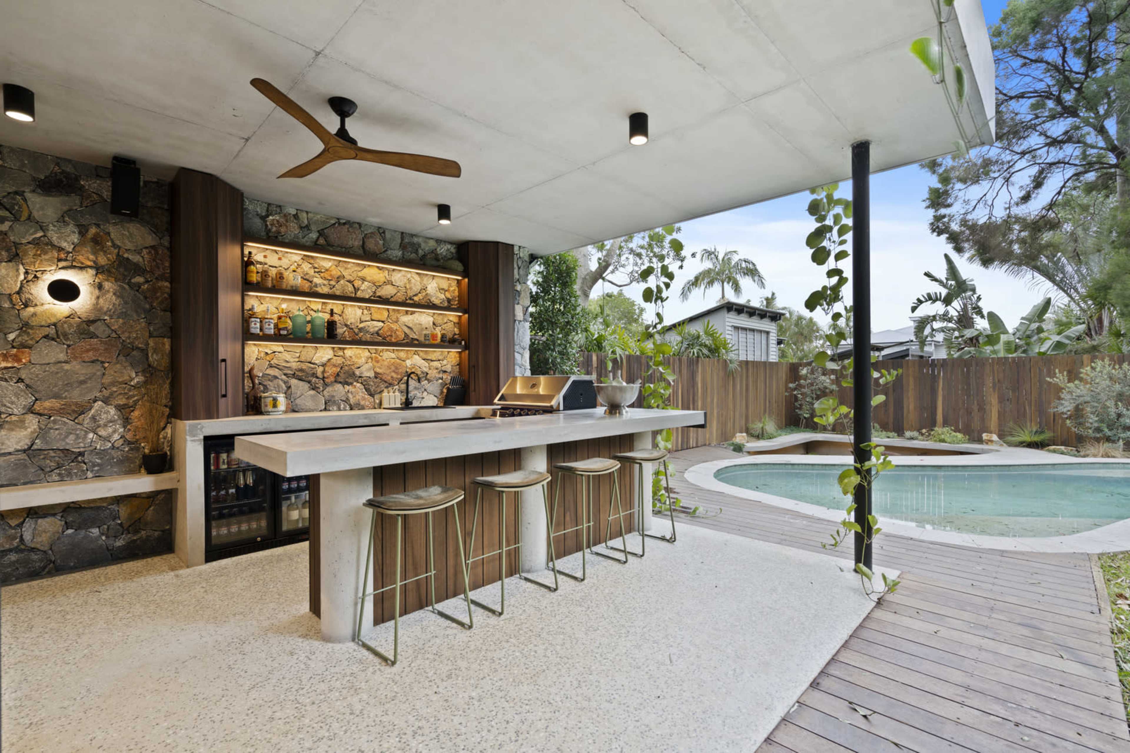 The image shows an outdoor bar area with a stone wall, a countertop, three stools, and a swimming pool surrounded by wooden decking and greenery.