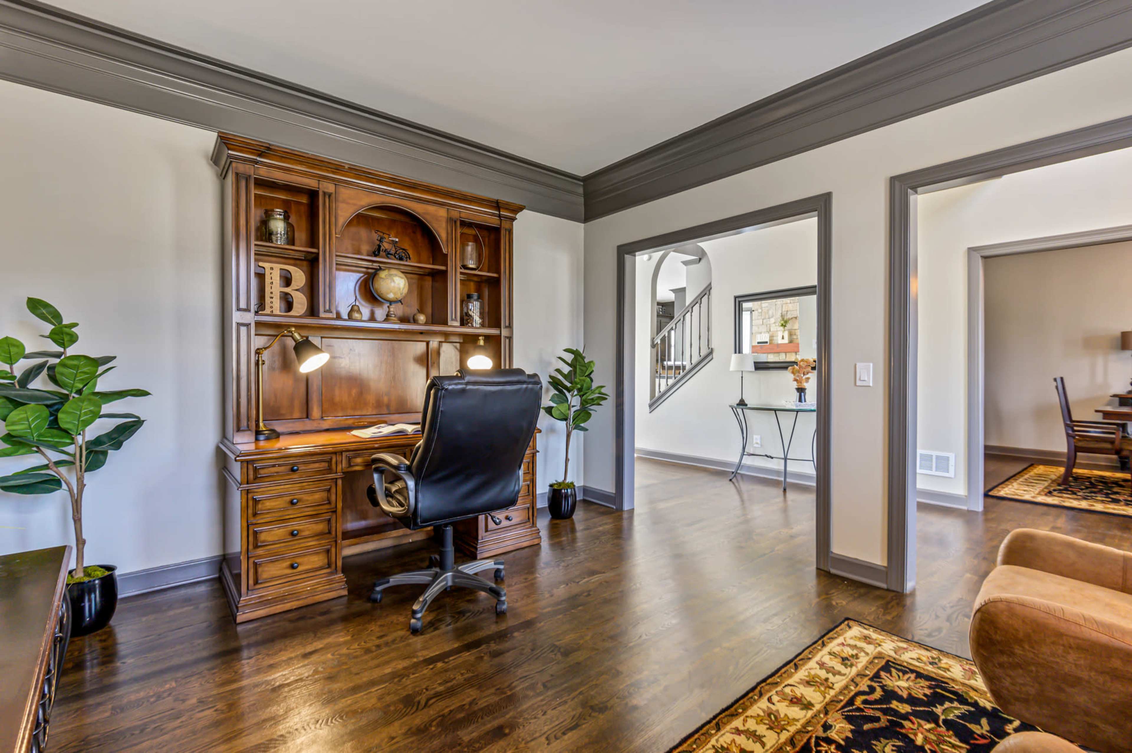 The image shows a home office with a wooden desk and chair, surrounded by potted plants and a view into an adjacent room.