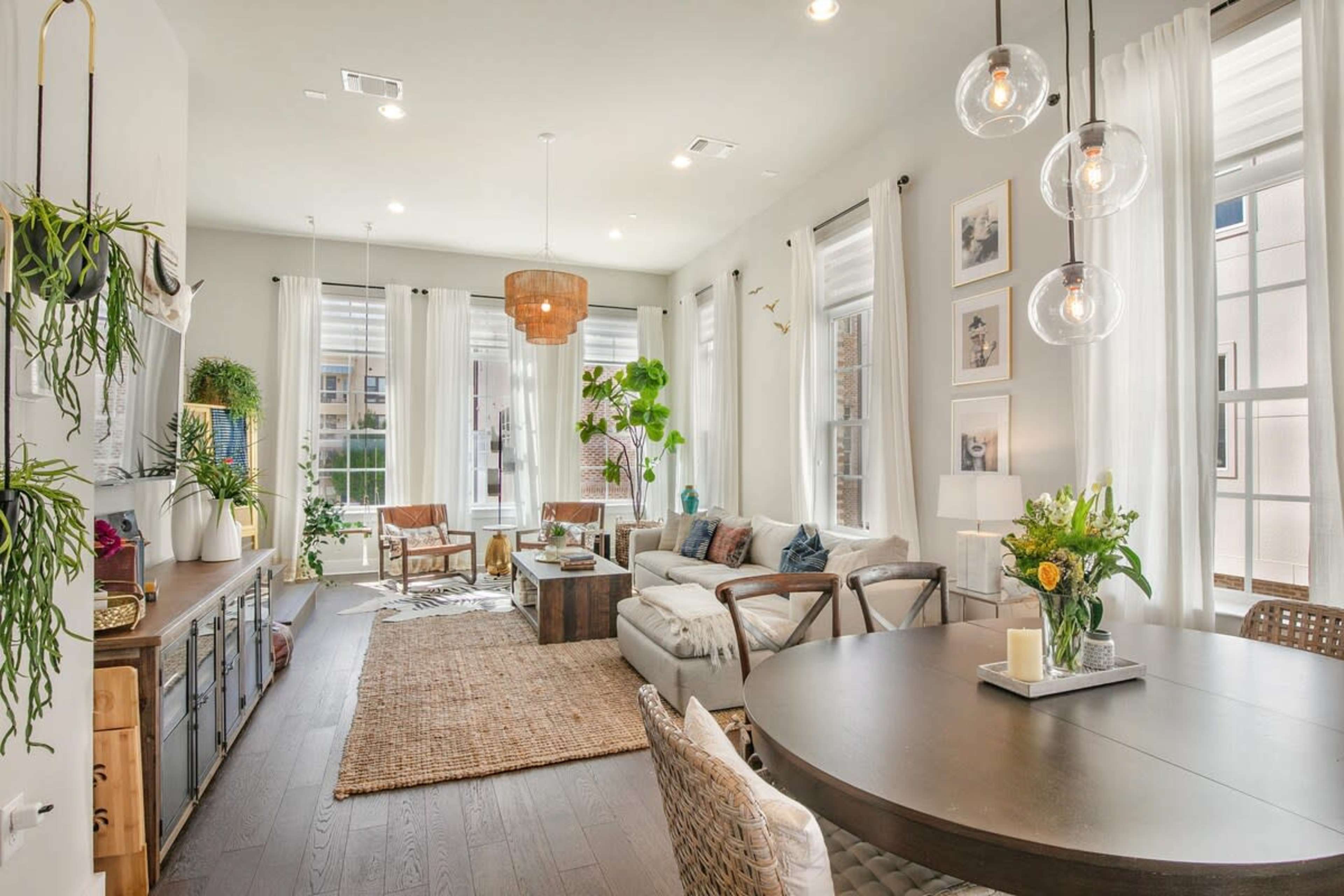 The image shows a bright living room with large windows, a comfortable sofa, a dining table, and decorative plants.