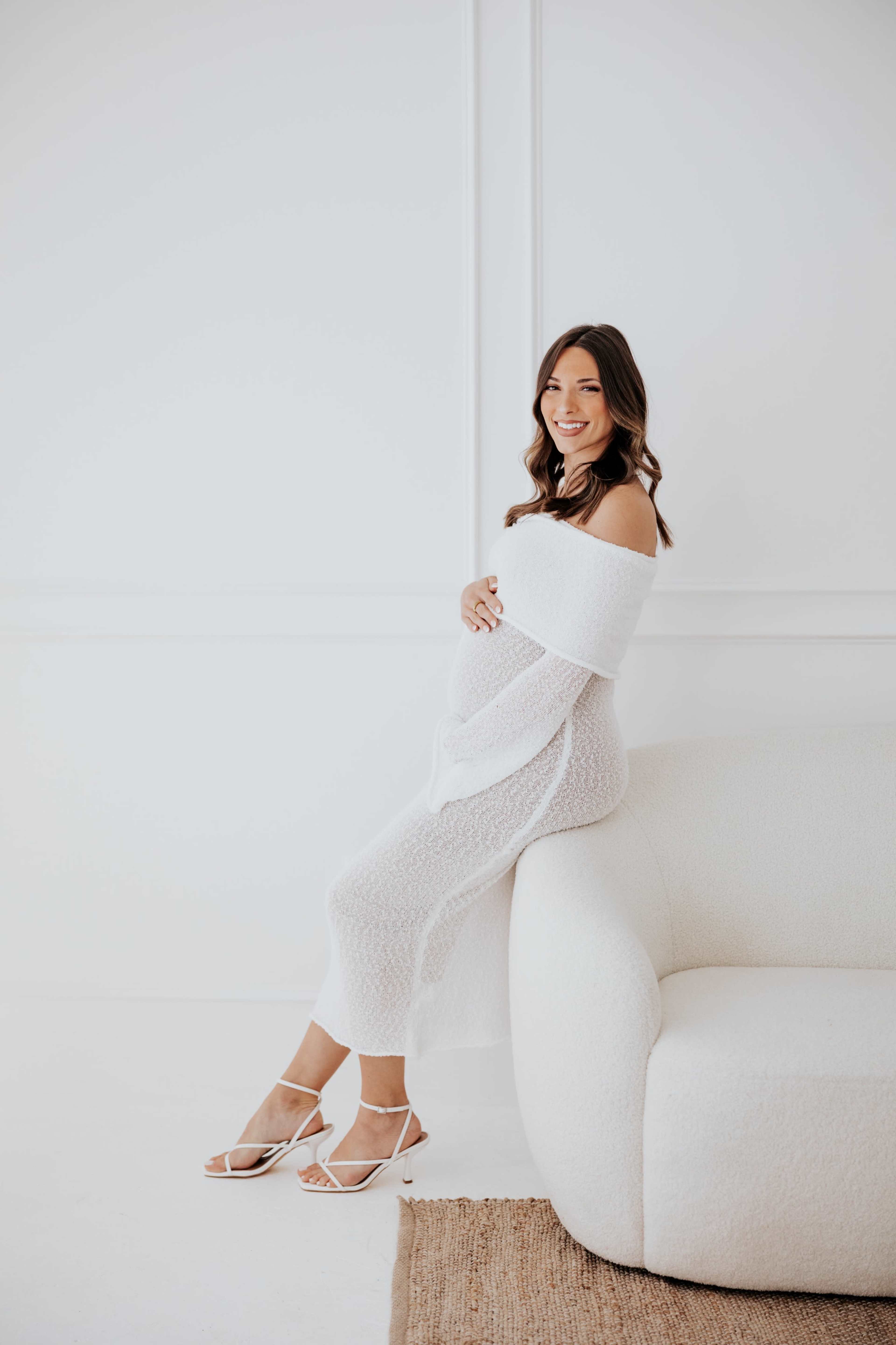 A pregnant woman in a white off-the-shoulder dress poses while leaning against a white textured sofa in a bright room.