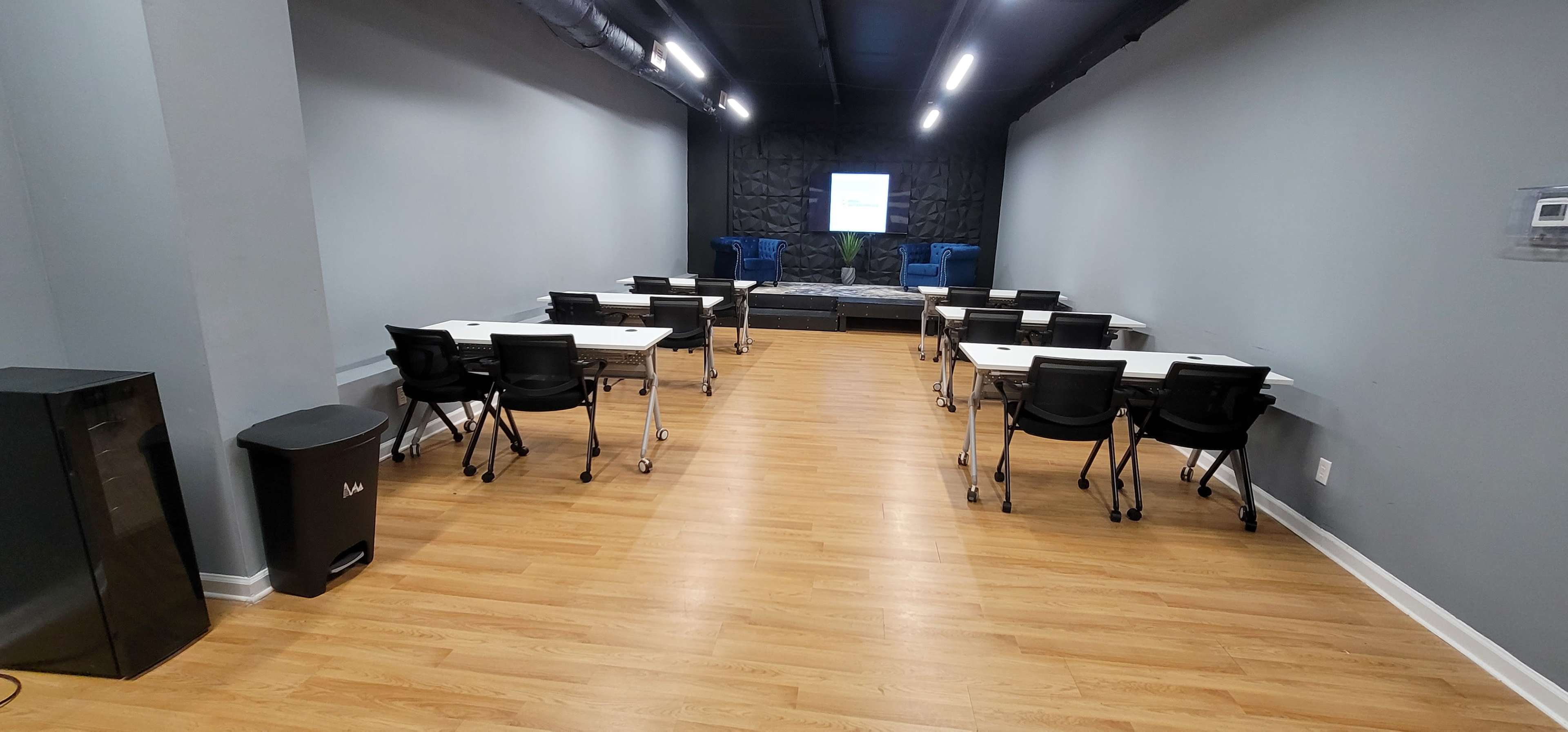 The room features several tables and chairs arranged in rows facing a screen at the front, with a seating area in the background.