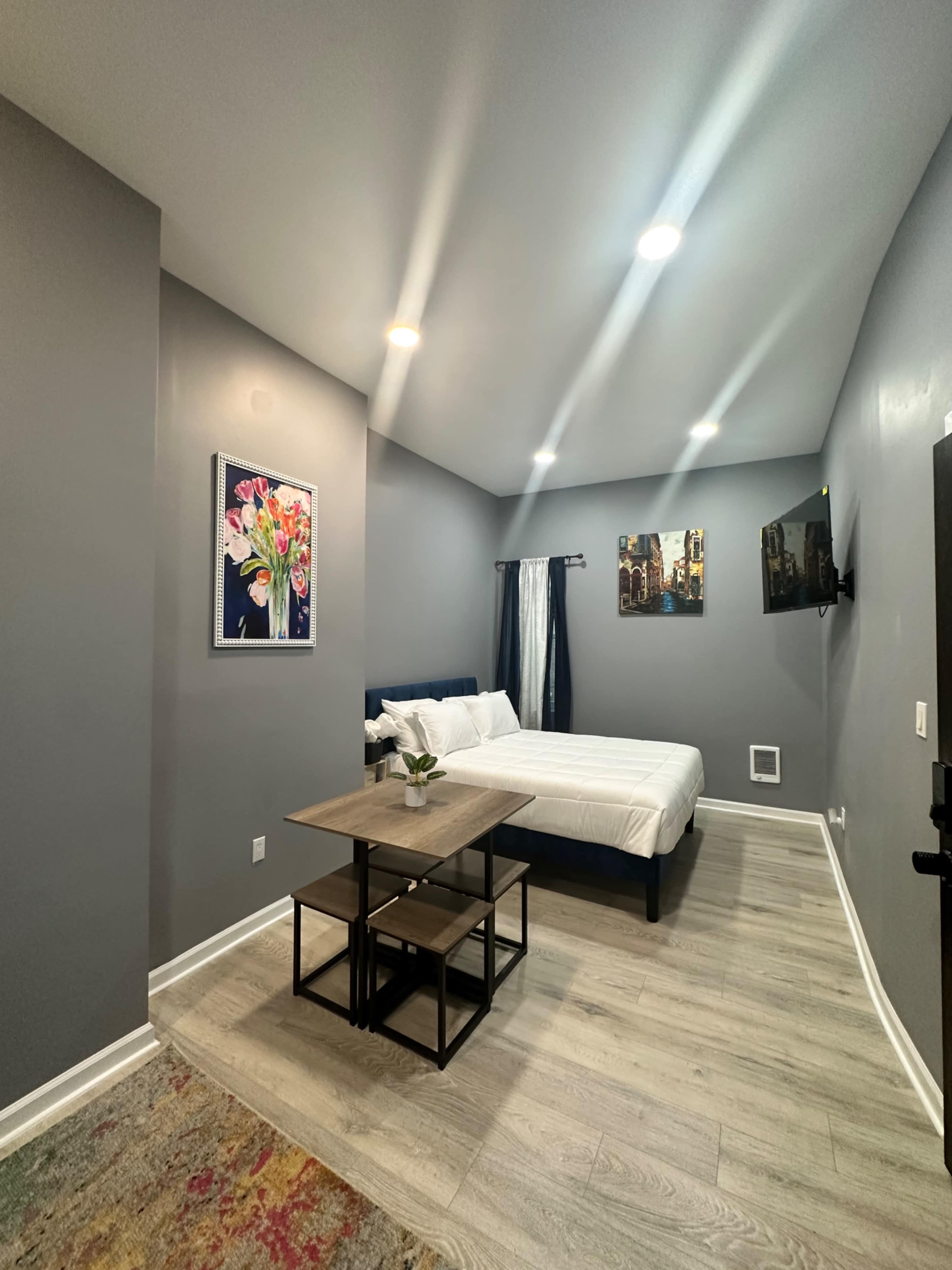 The image shows a modern bedroom with a bed, a wooden table, and artwork on the walls, featuring gray walls and a light-colored floor.