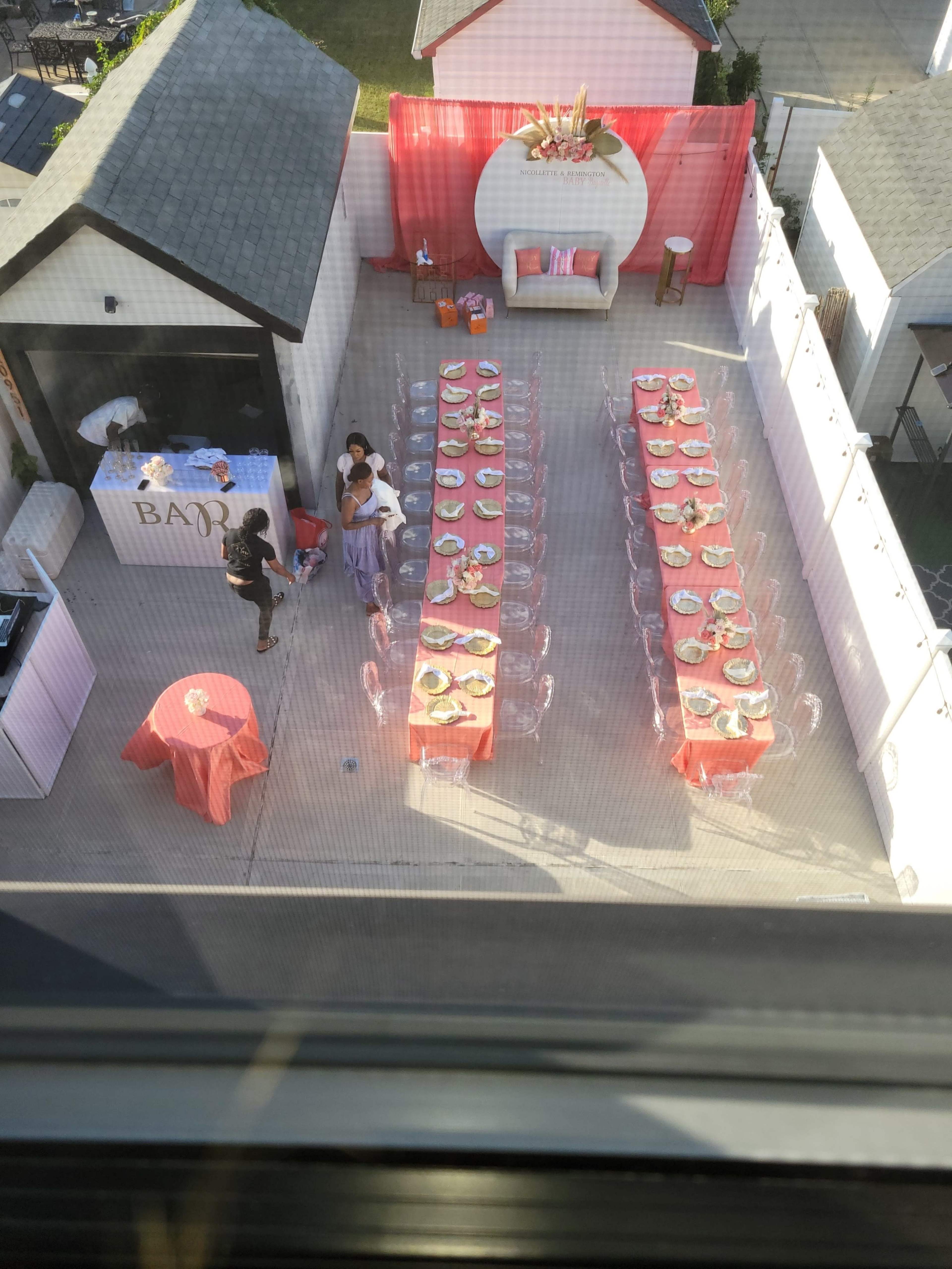 An outdoor event setup with two long tables arranged with plates and decorations, surrounded by a white wall and a kitchen area in the background.