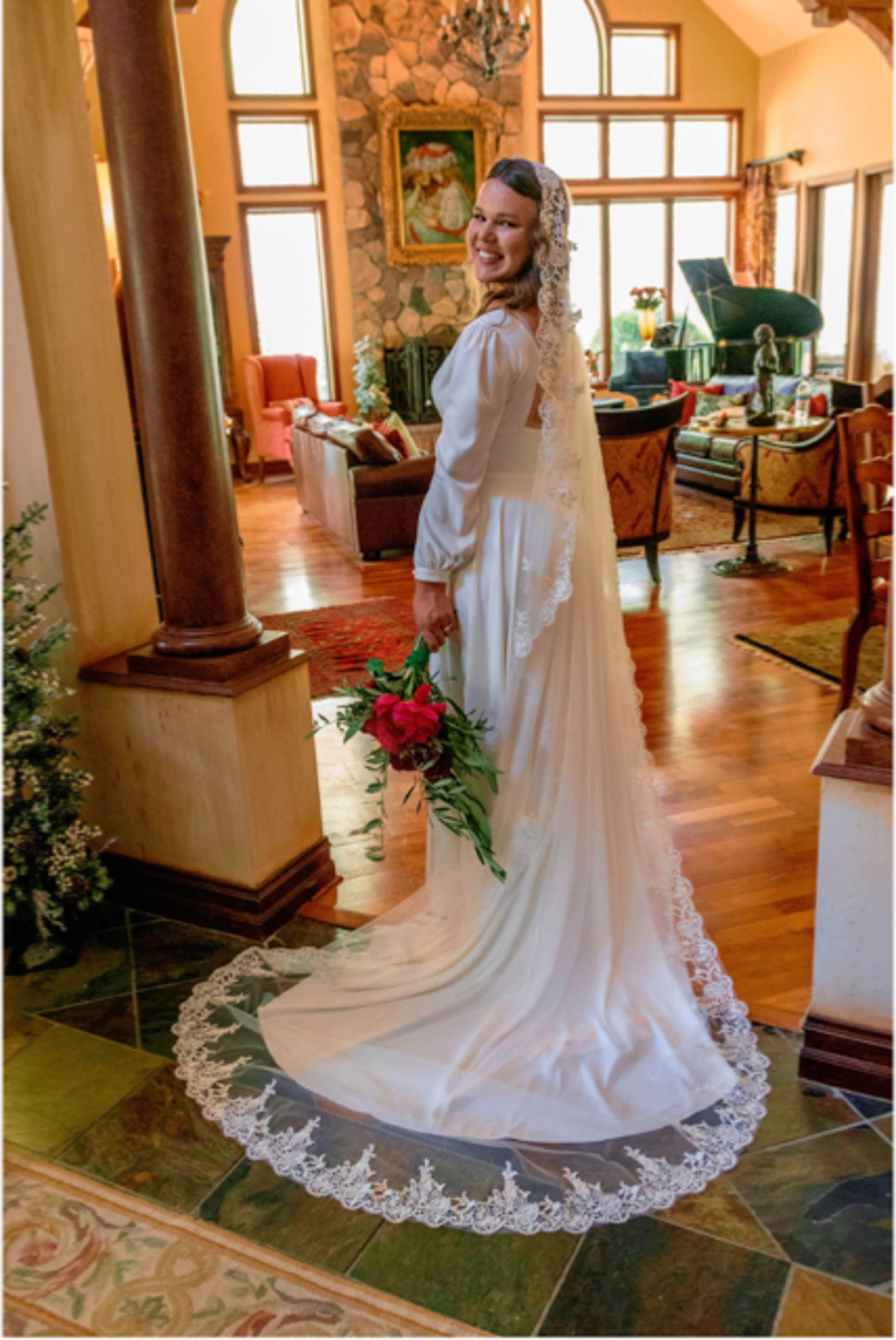 A bride in a flowing white dress and veil holds a bouquet of red roses as she poses in a beautifully decorated living room.