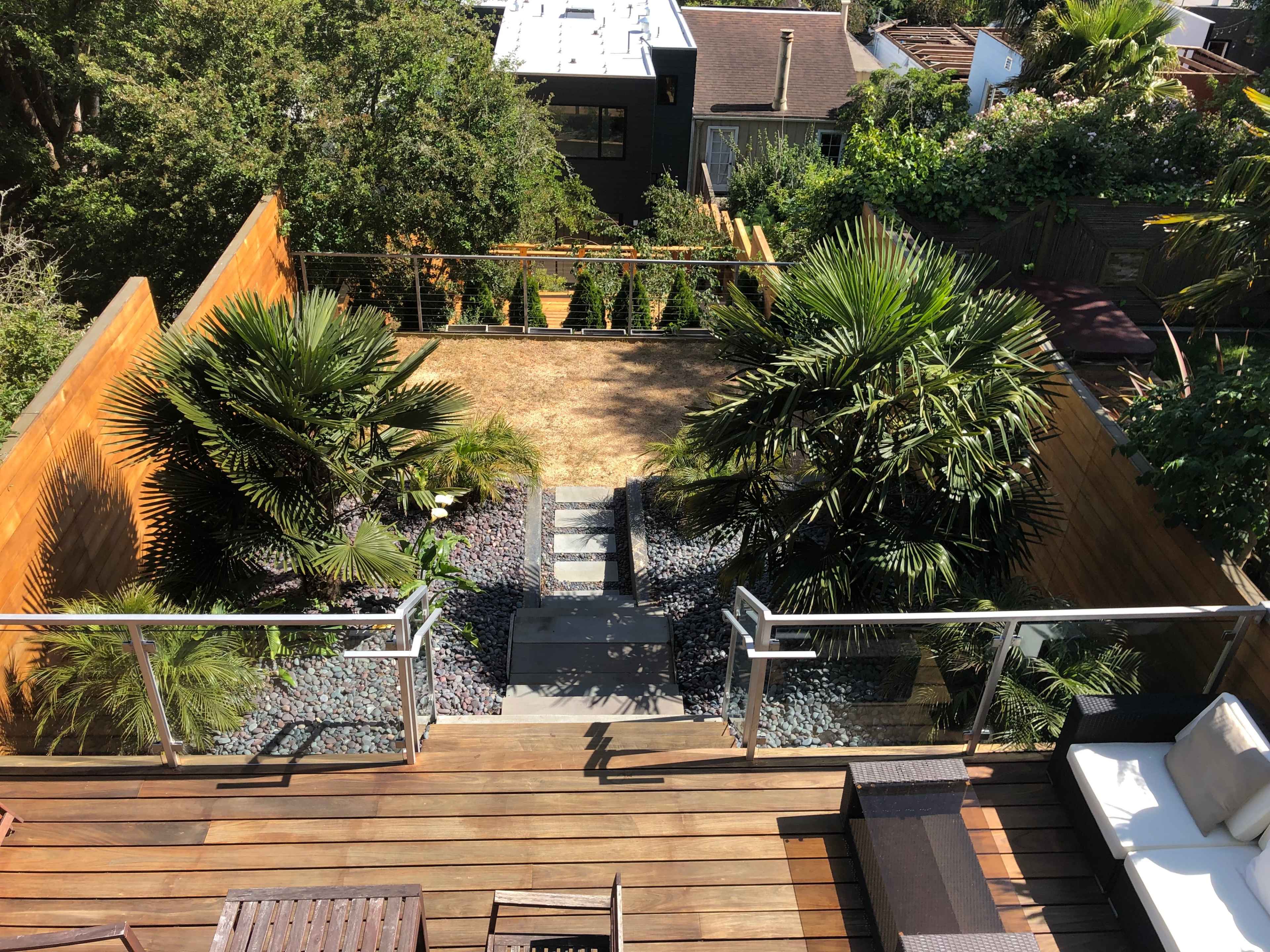 A backyard with a stone pathway leading down to an area featuring two palm trees and gravel, enclosed by wooden fencing.