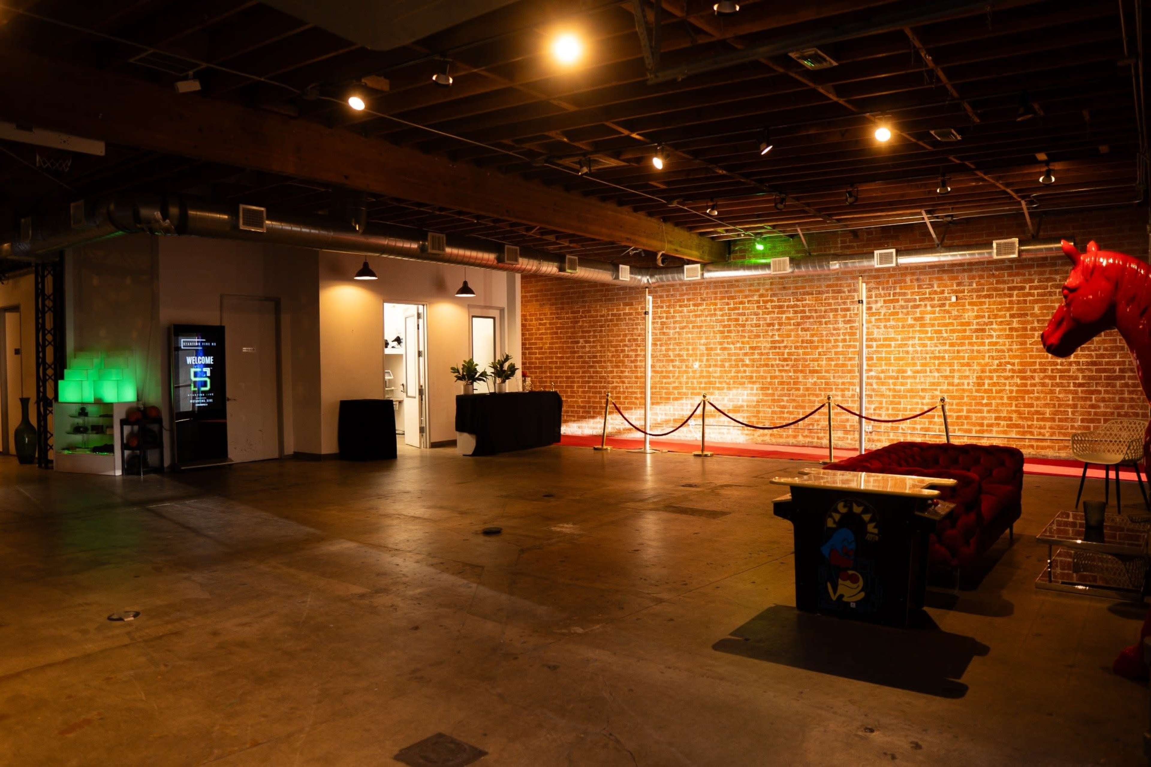 The image shows an industrial-style interior with exposed brick walls, a red sofa, a vending machine, and a red horse statue in a spacious, well-lit area.
