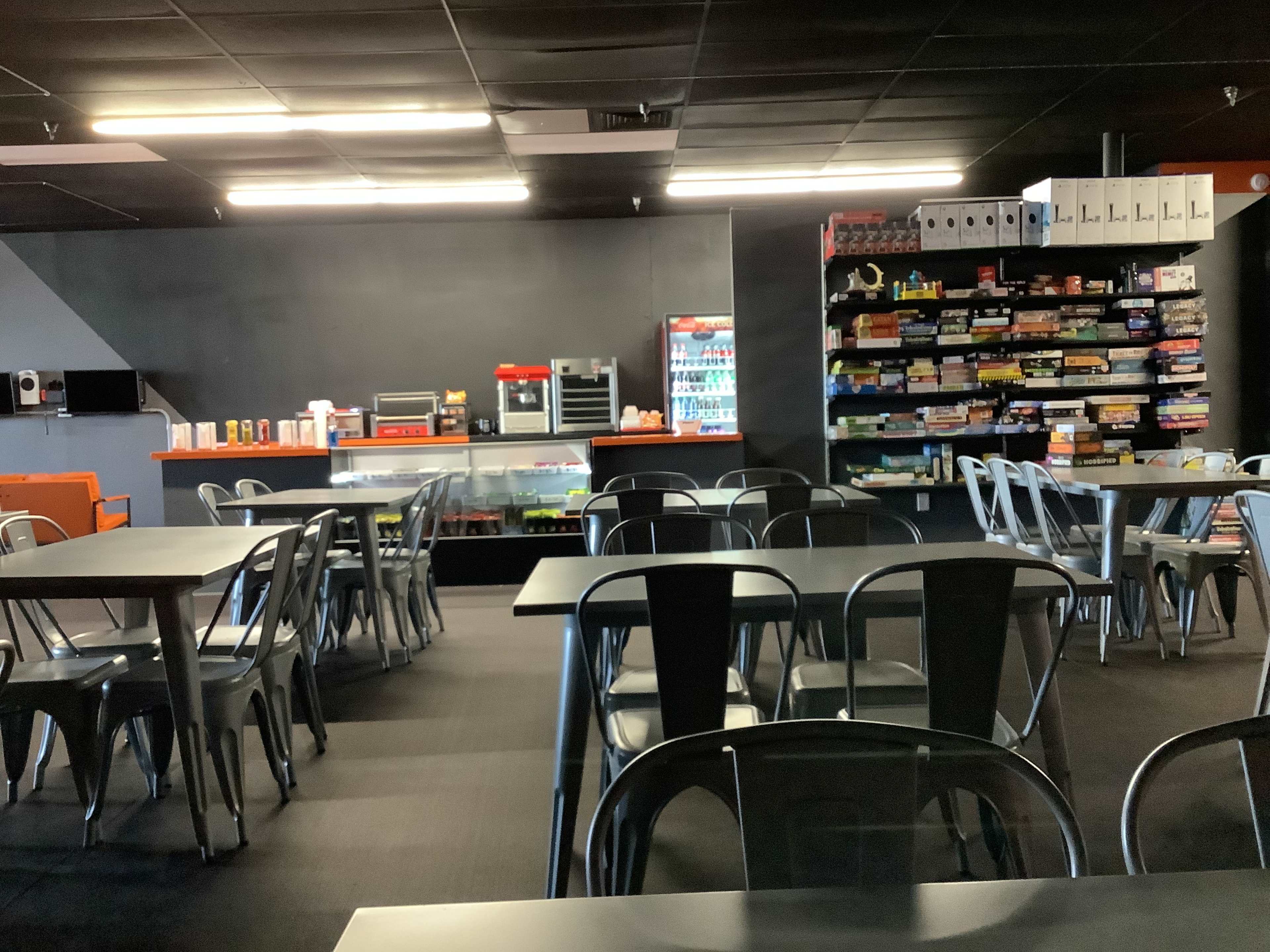 The image shows a dimly lit café or game room with metal tables and chairs, a snack bar with drinks, and shelves stocked with board games.