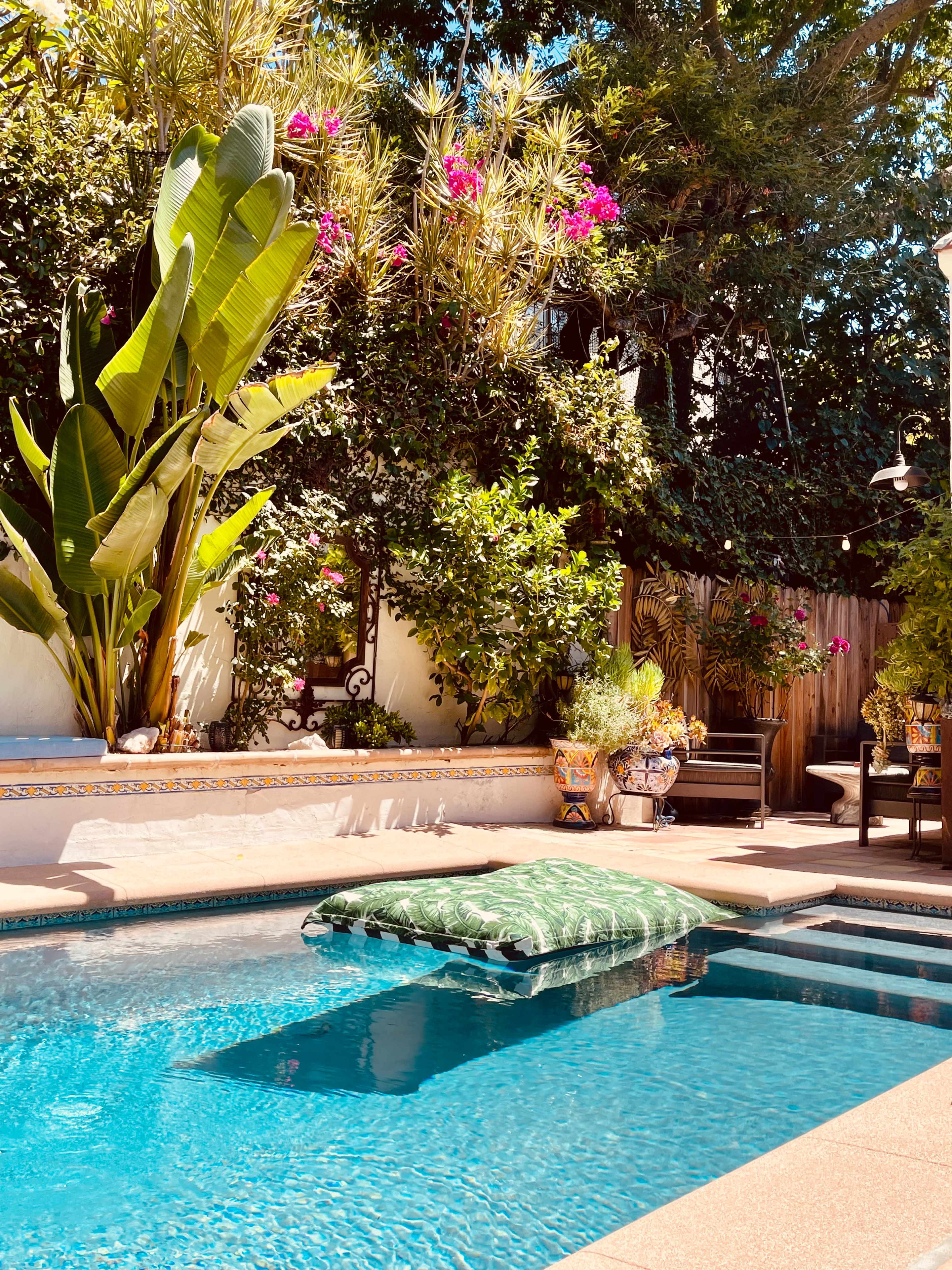 A swimming pool features a large green float surrounded by tropical plants and colorful potted flowers.