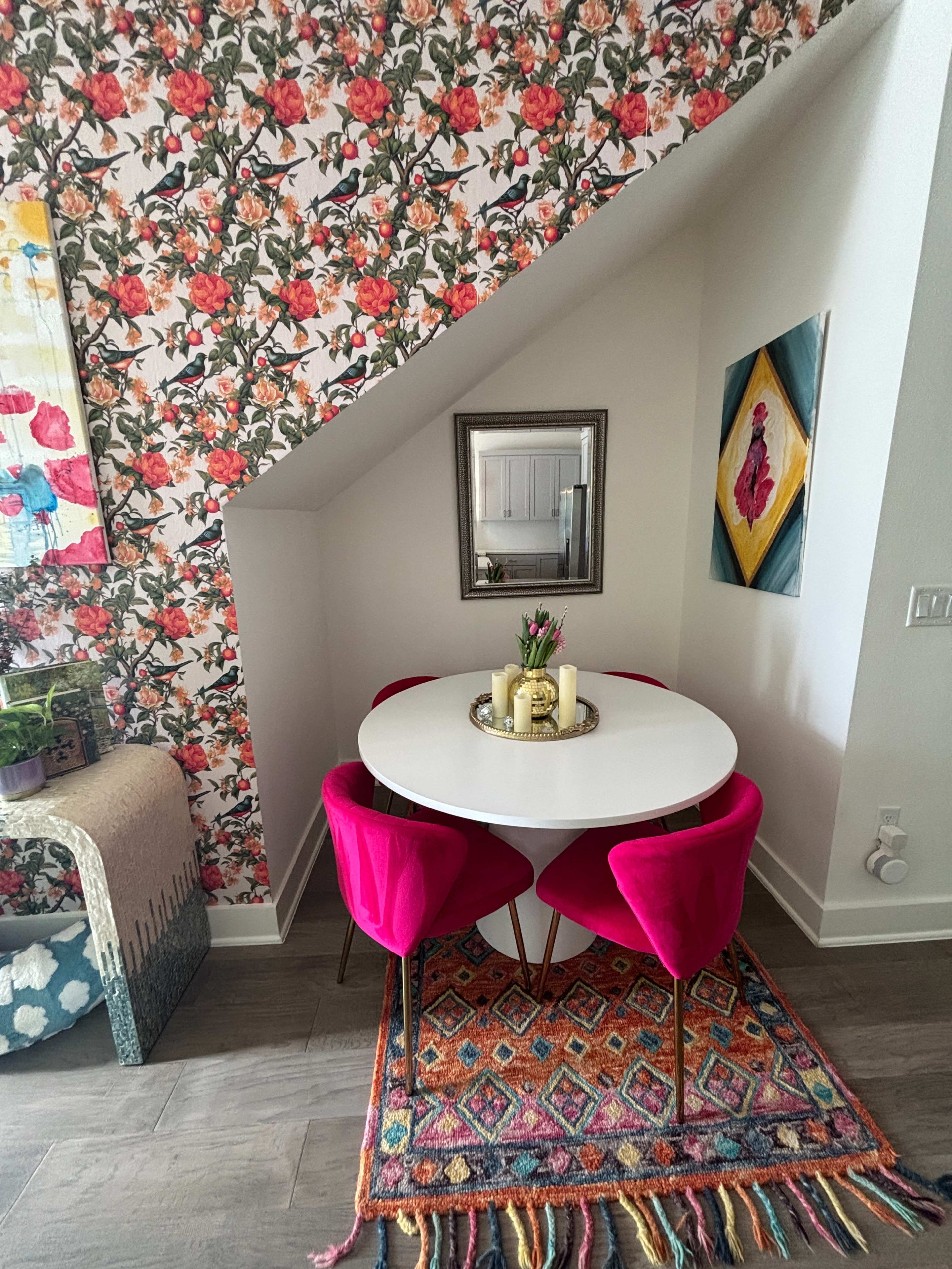 A small dining area features a round white table surrounded by four pink chairs, positioned against a floral wallpaper and illuminated by natural light.