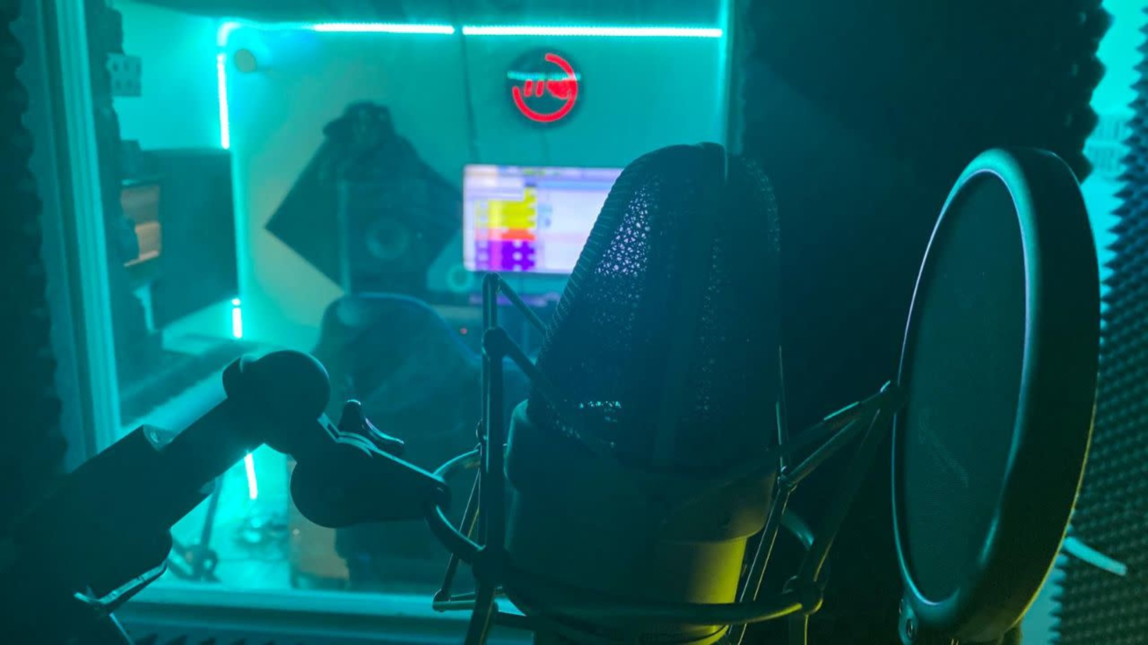 A recording studio booth with a microphone in the foreground and colorful LED lights illuminating the background equipment.