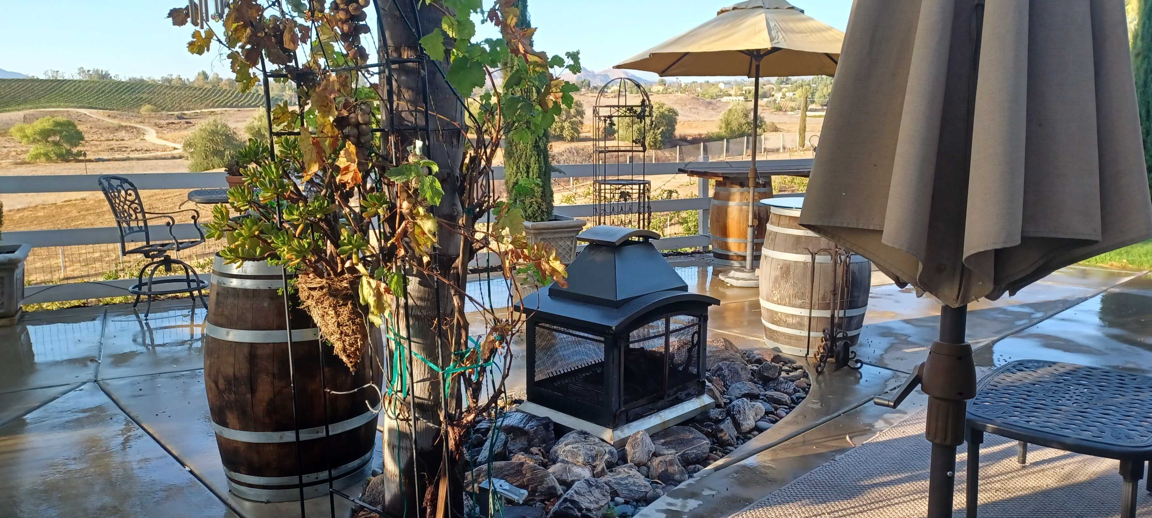 The image shows a patio area with a metal fire pit surrounded by rocks, wooden barrels, and a large umbrella, set against a scenic outdoor landscape.
