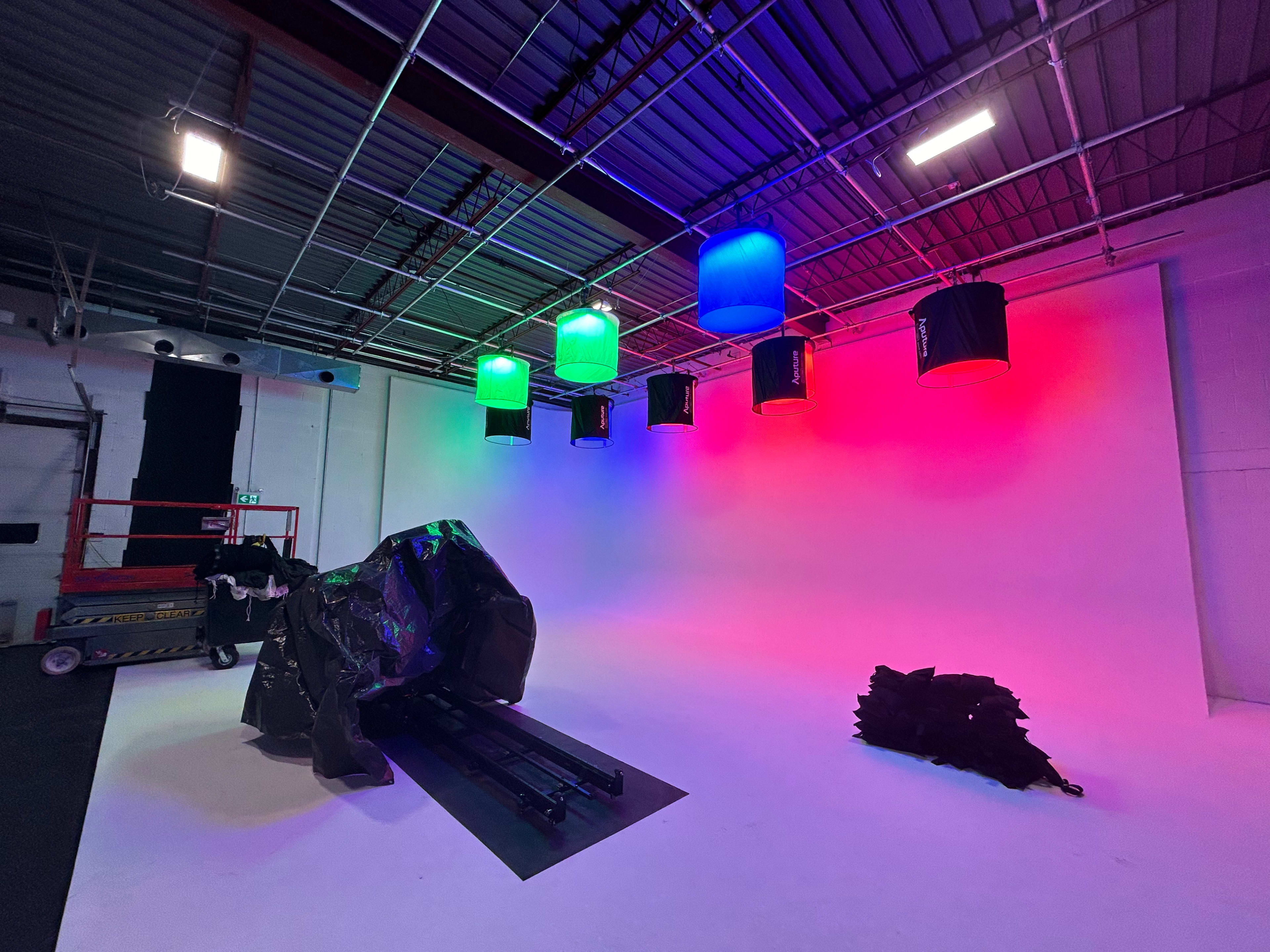 A spacious studio with a white backdrop and colorful lights hanging from the ceiling, along with equipment and tarps on the floor.