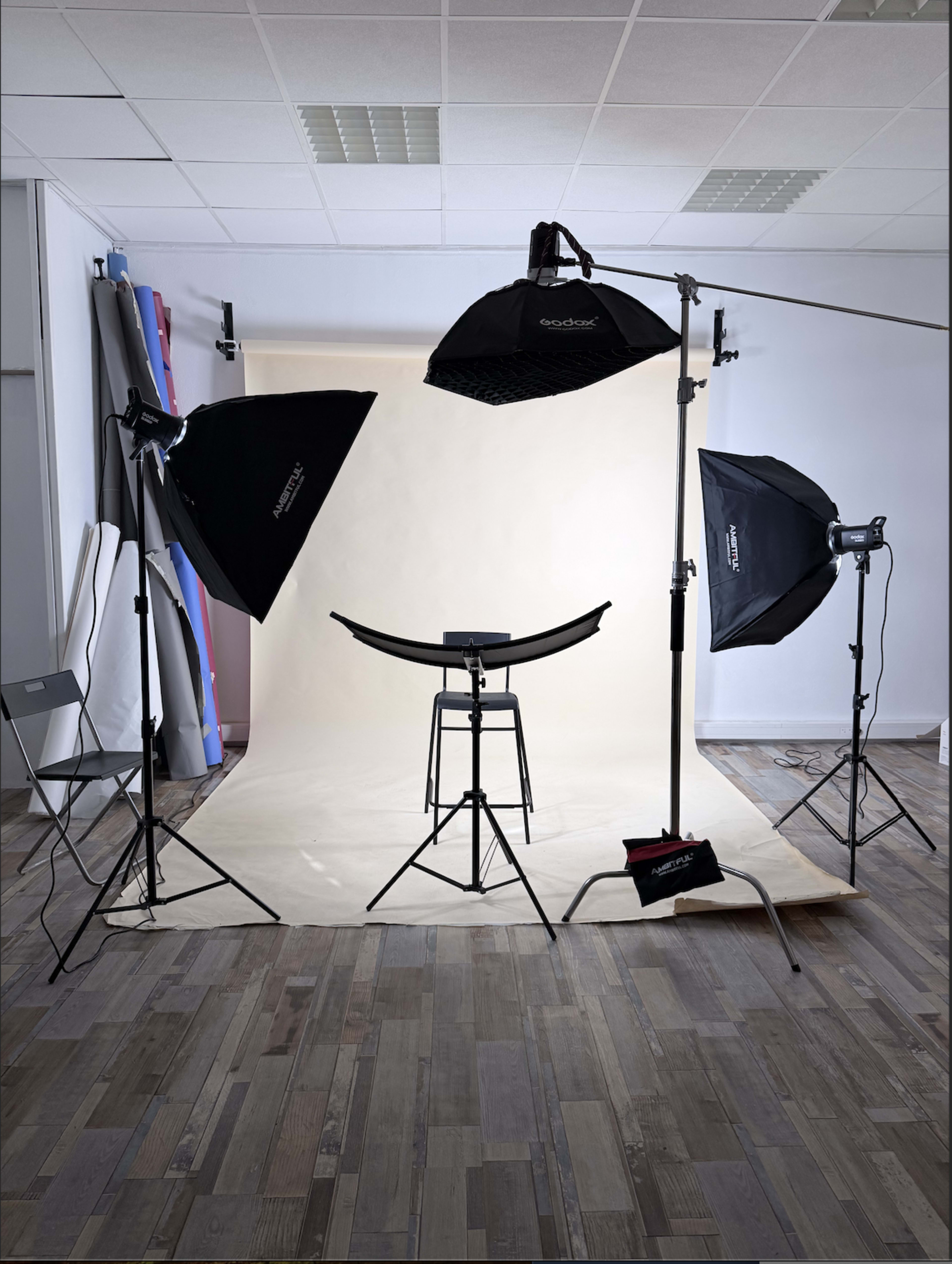 The image shows a photography studio setup with multiple light sources, a curved backdrop, and a stool positioned in front of a beige background.