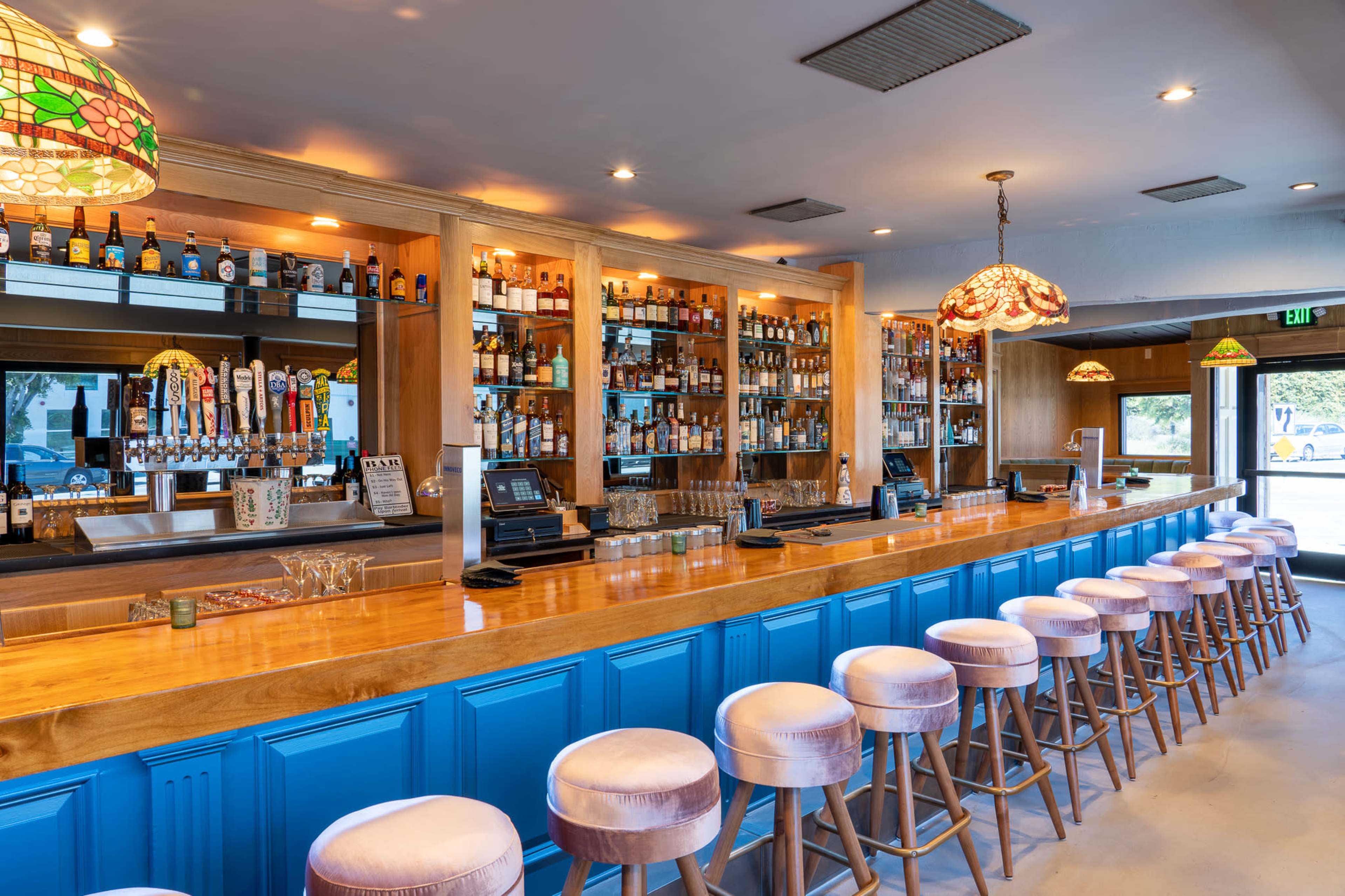 A brightly lit bar features a polished wooden countertop, shelves filled with various liquor bottles, and a row of upholstered stools.