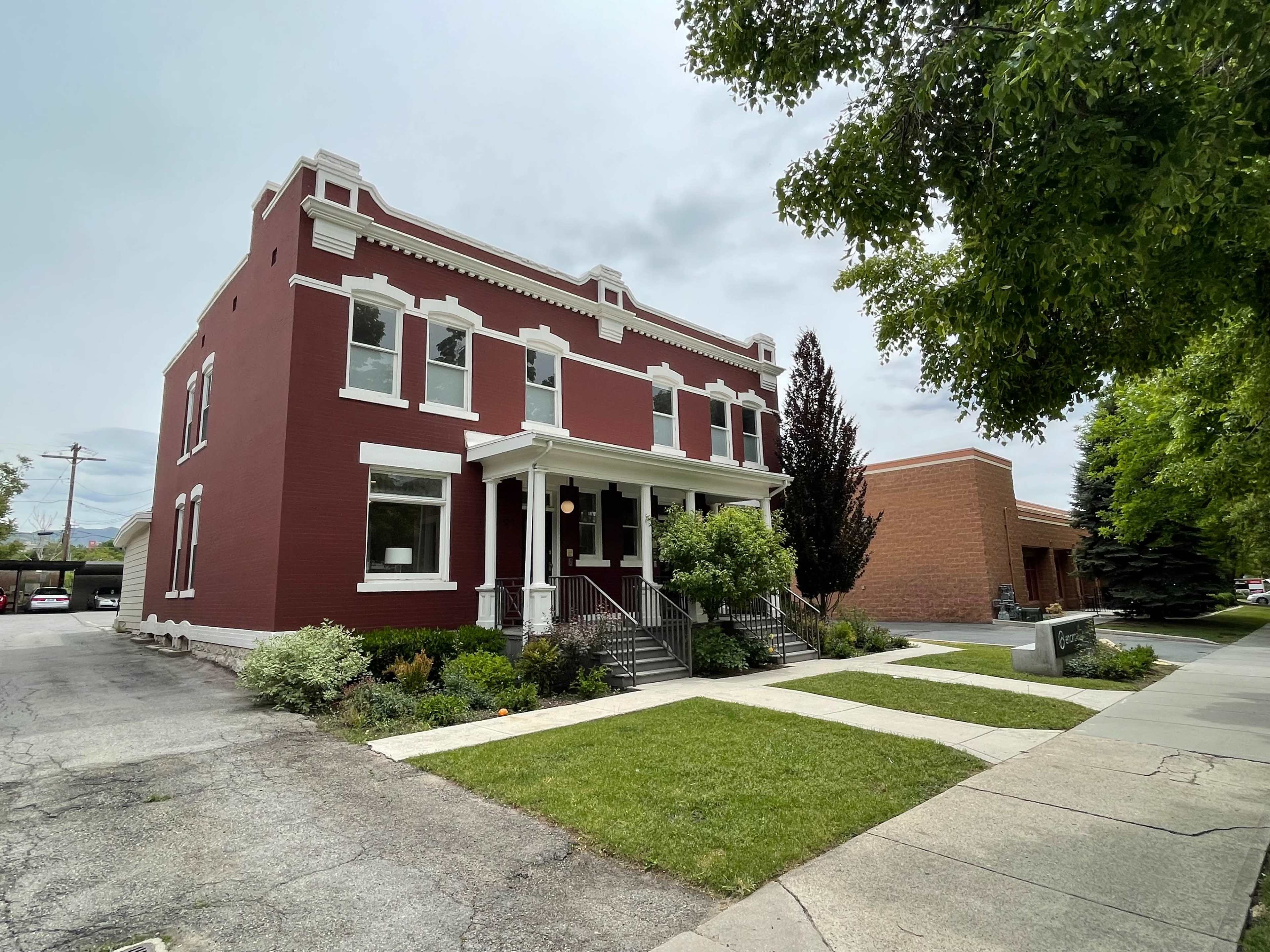 A red-brick building with white trim, featuring a front porch and landscaped greenery, situated on a sidewalk beside a paved road.