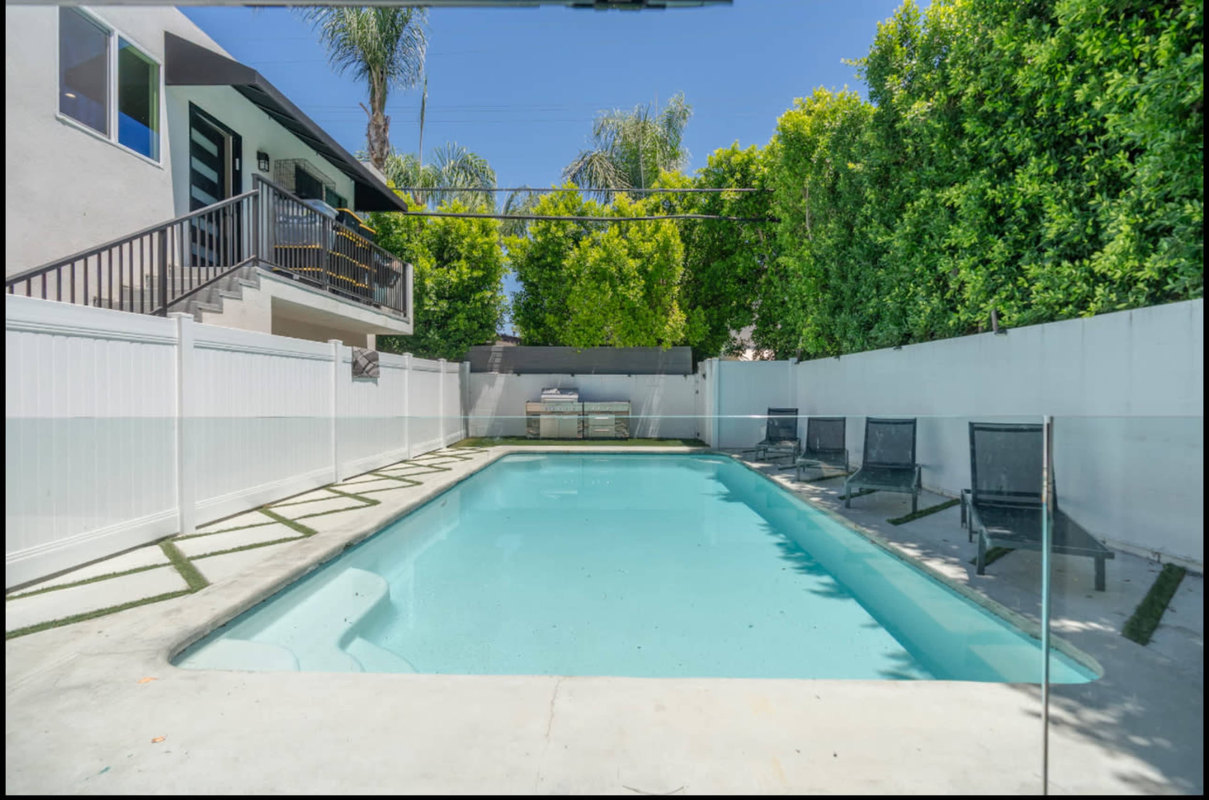 The image shows a backyard with a rectangular swimming pool surrounded by a concrete patio and black lounge chairs, bordered by a white fence and greenery.